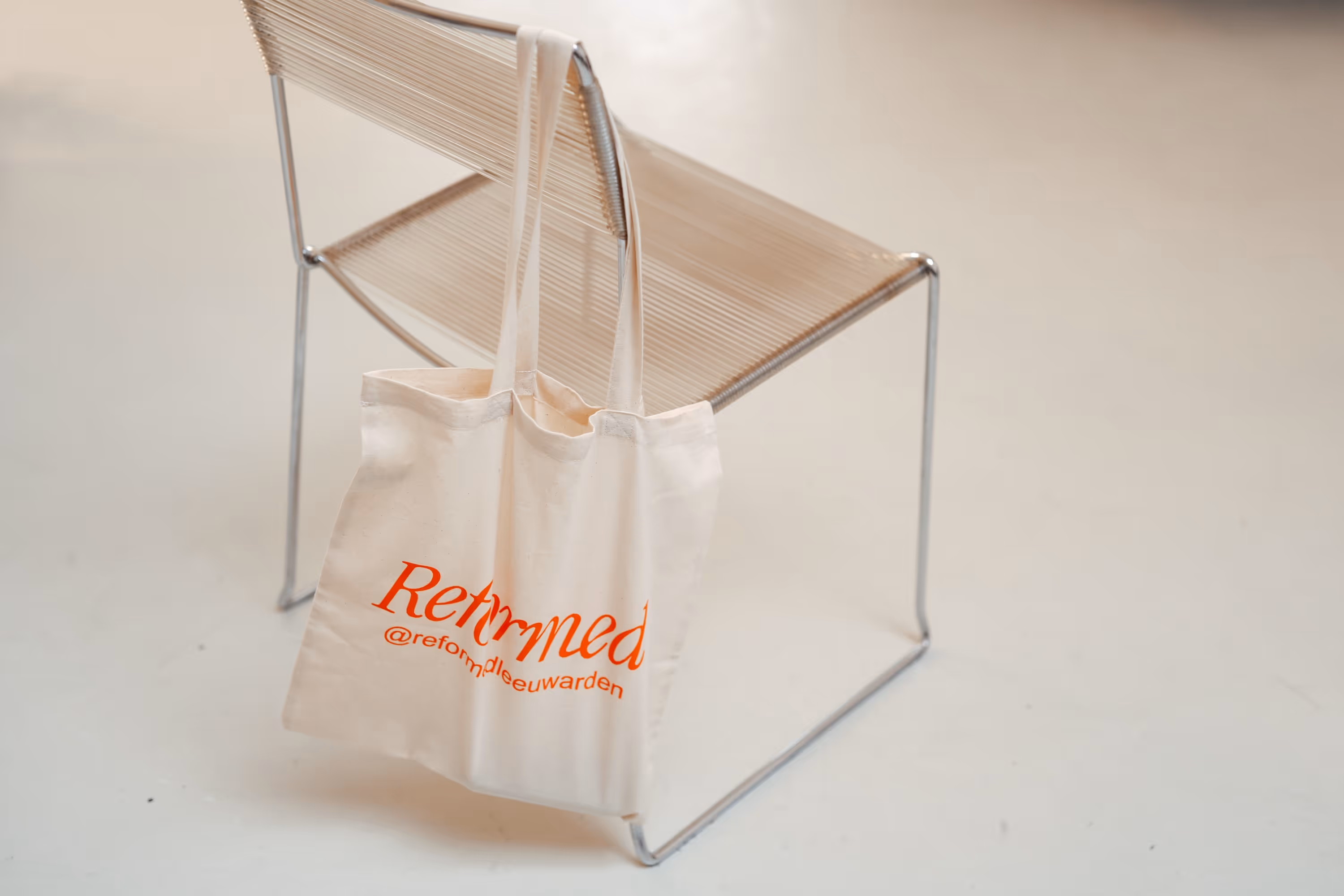 Crèmekleurige stoffen draagtas met rode tekst 'Reformed @reform_leeuwarden' hangt aan een minimalistische metalen stoel met geweven rugleuning.