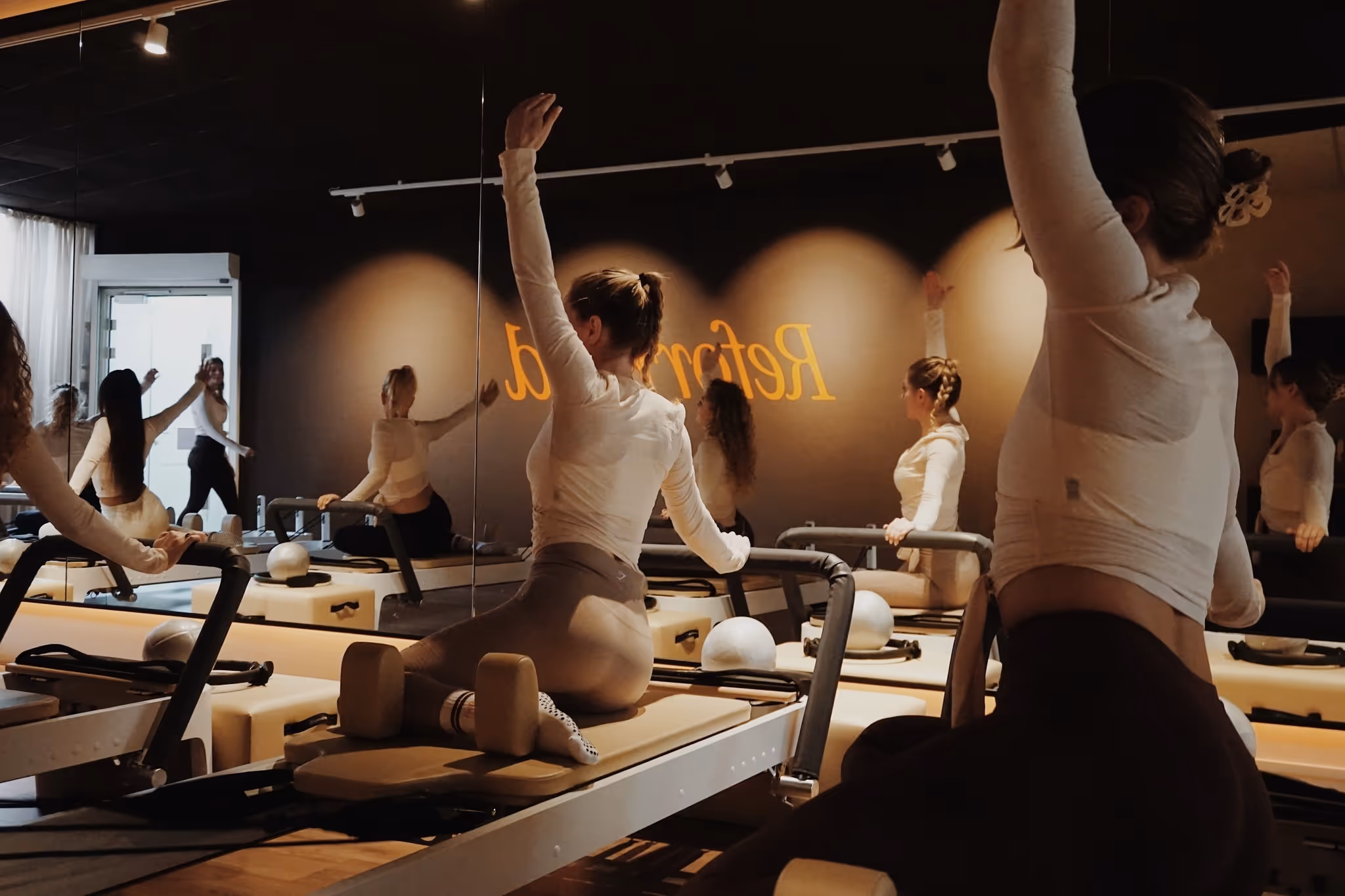 Vrouwen doen Pilatesoefeningen op reformertoestellen in een studio met spiegels en gedimde verlichting.