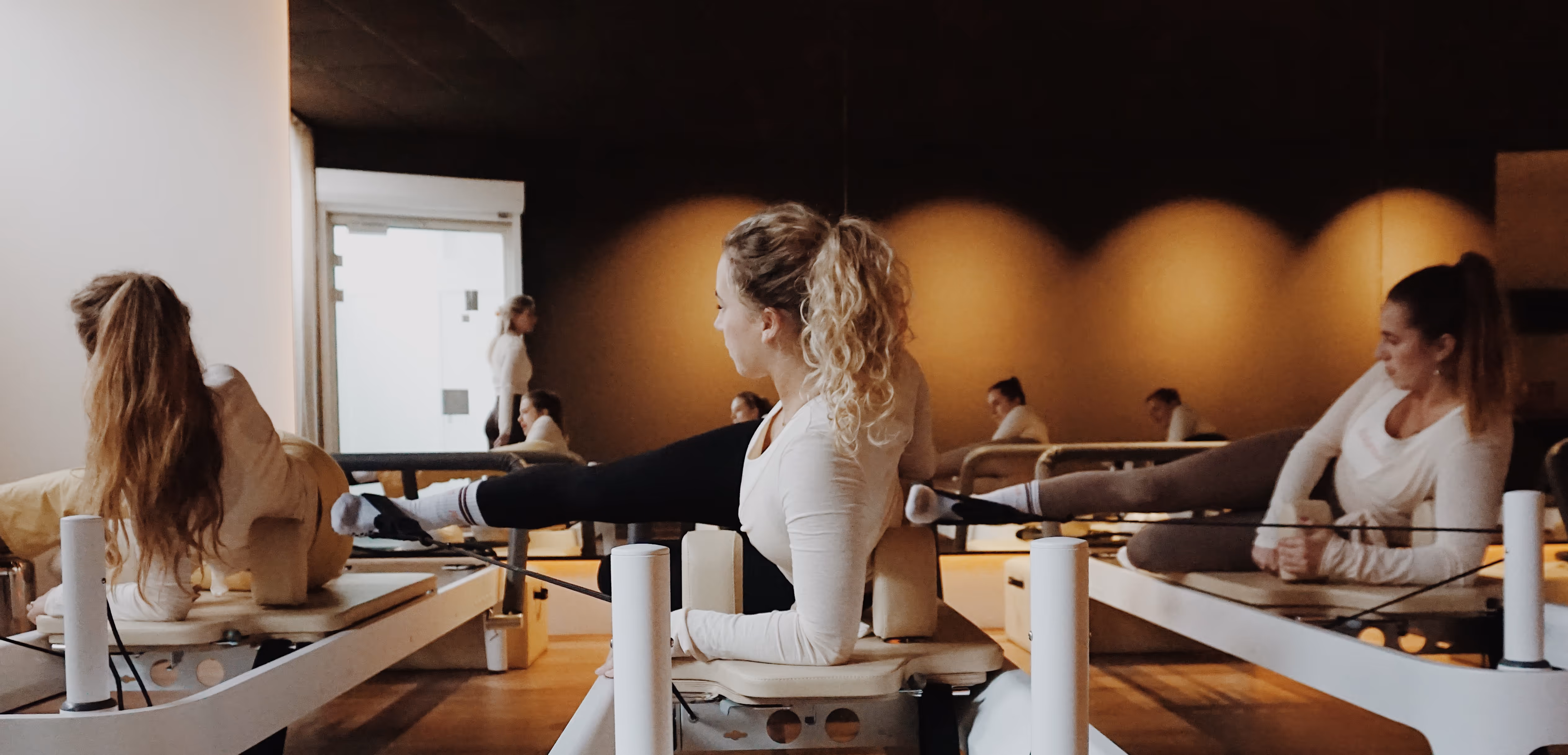 Drie vrouwen doen oefeningen op pilatesapparaten in een studio met warme verlichting.