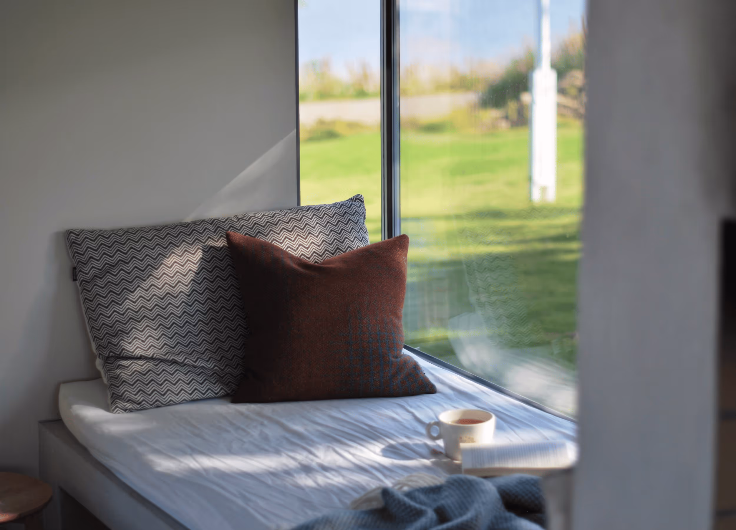 Behagelig vindussofakrok med puter og utsikt over gress og natur på Skagenhaugen – ro og komfort i nord-norge.