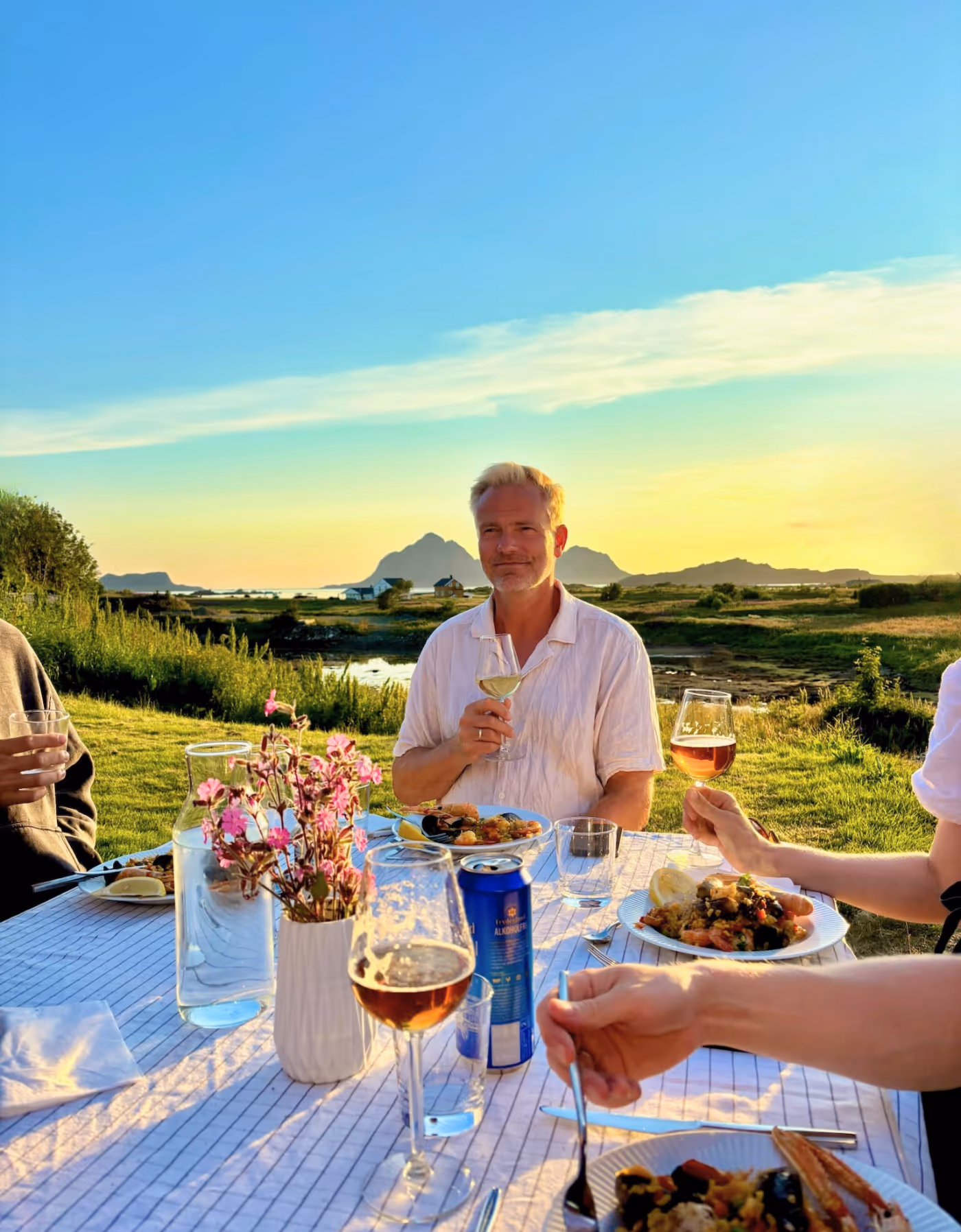 En mann sitter rundt et bord med et smil om munnen, omgitt av familie utendørs i Vesterålen, utenfor Skagenhaugen