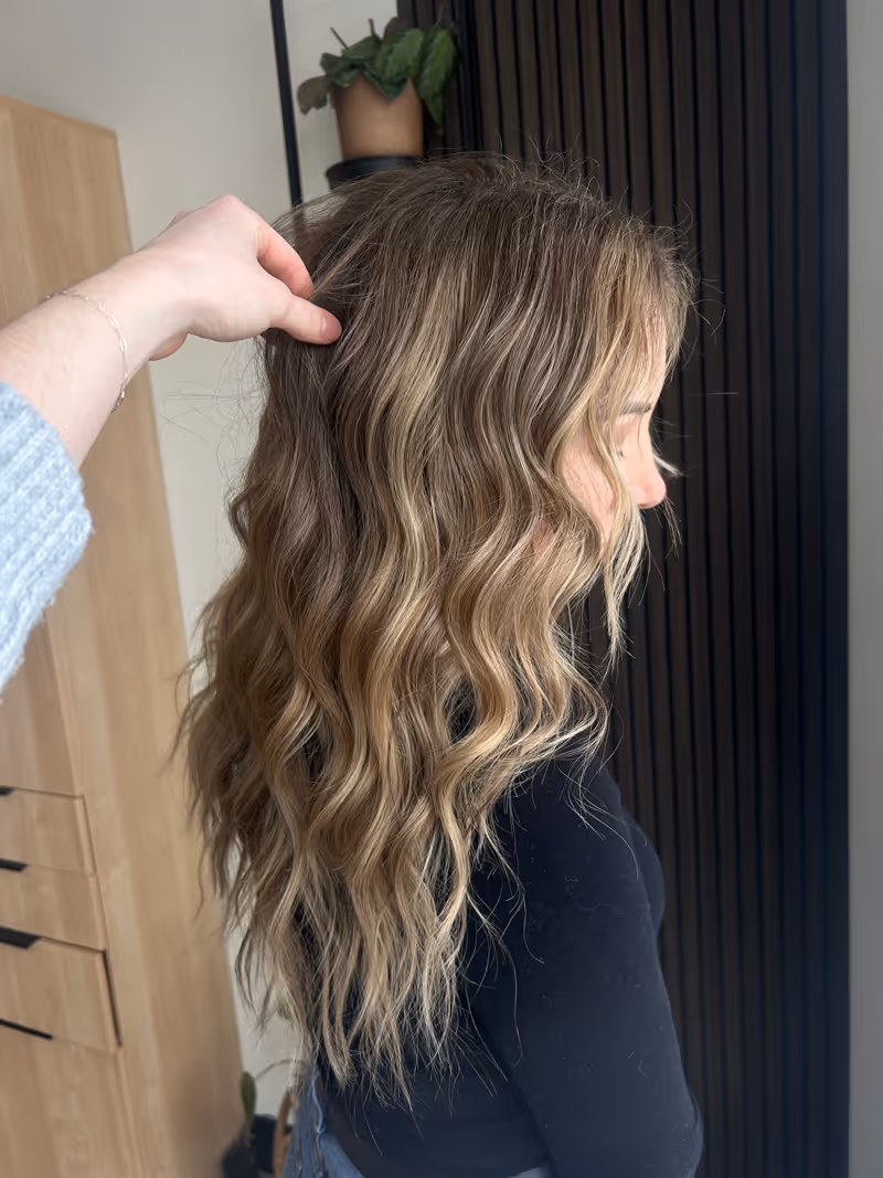 Side profile of a woman with long, wavy, highlighted blonde hair being styled by a hand.