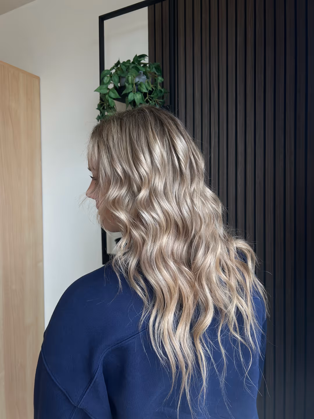 Person with long, wavy blonde hair wearing a dark blue top, viewed from the back and side.