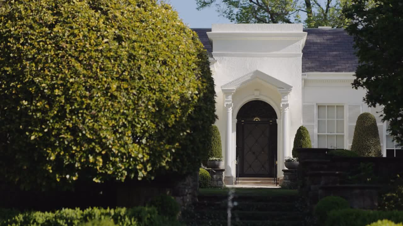 Elegant white estate entrance with black door framed by manicured hedges and topiary trees.