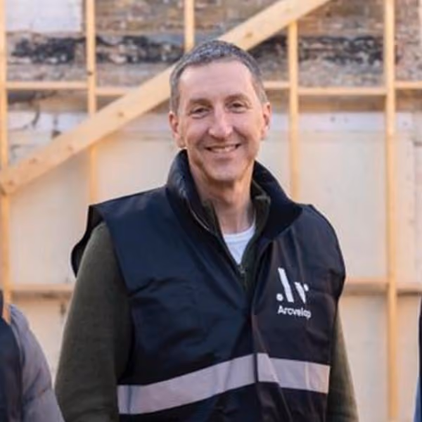 Smiling man wearing a black Arcoverde vest and green long-sleeve shirt, standing in front of a wooden frame.