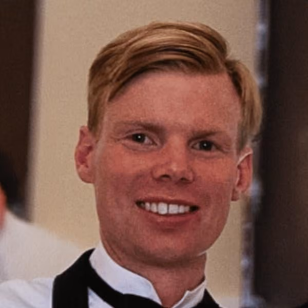 Smiling man with blonde hair wearing a white shirt and black bow tie in an indoor setting.