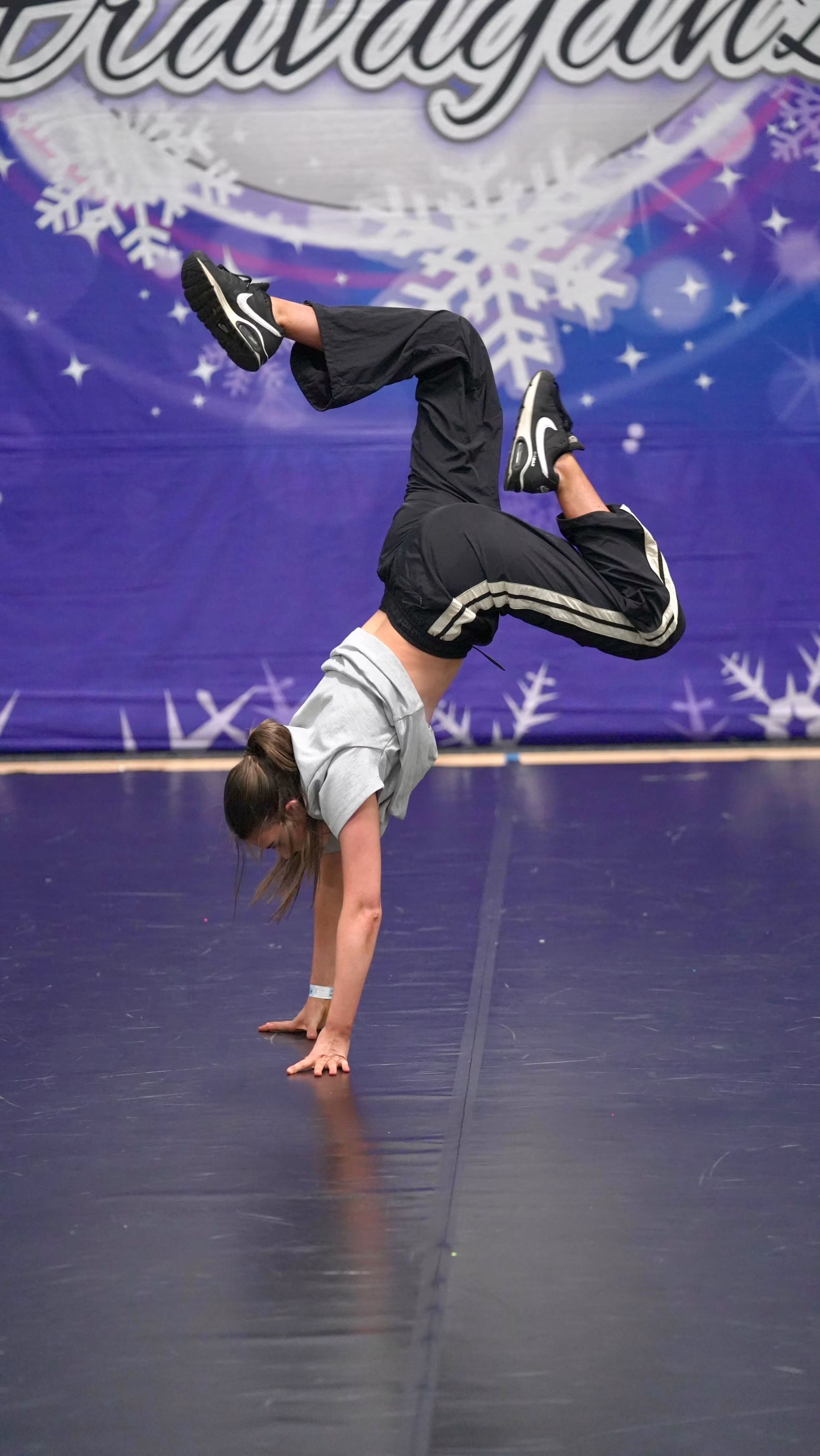 Young female dancer balancing on hands with bent legs on a stage with a purple snowflake backdrop.