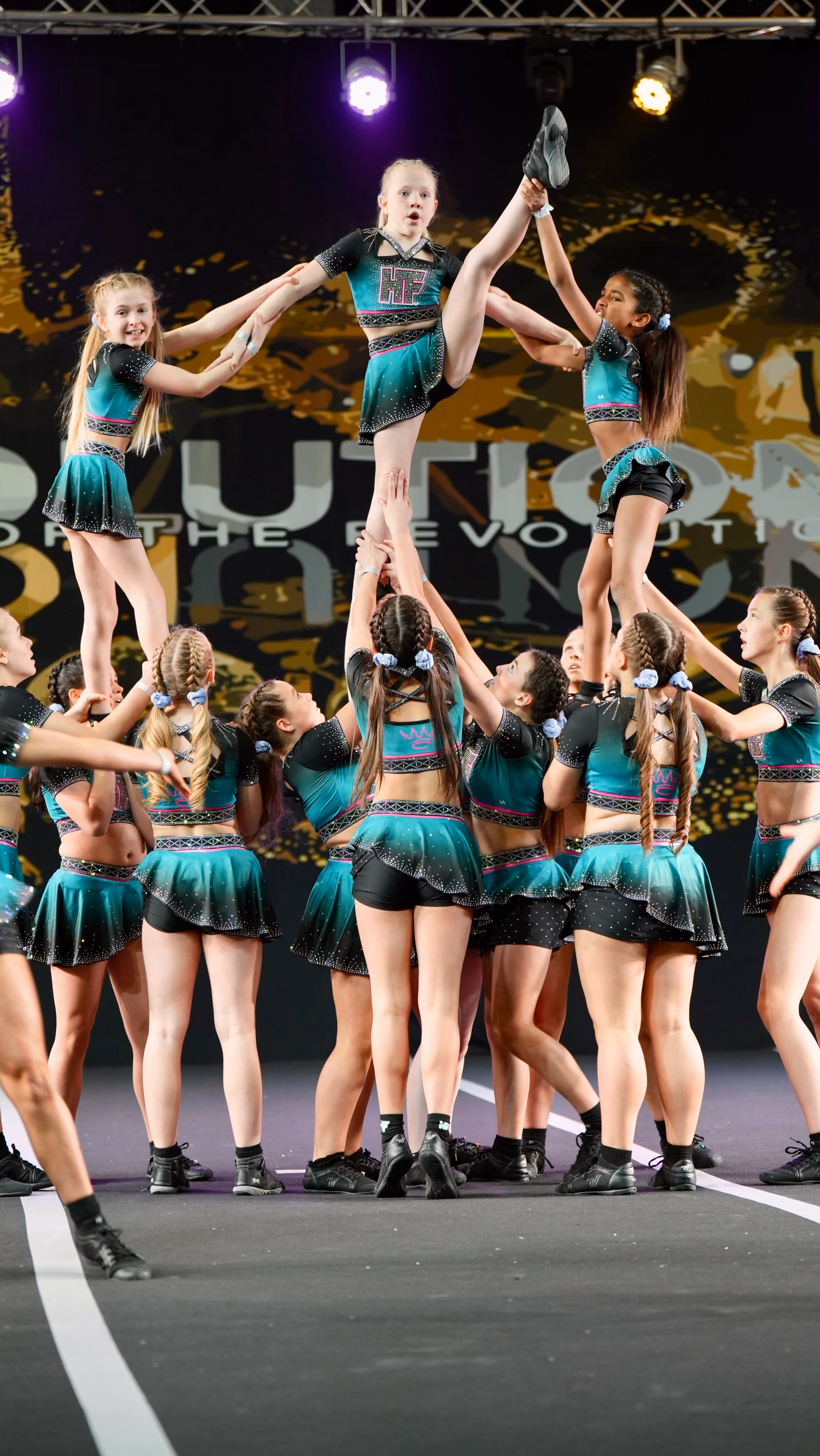 Cheerleading team lifting two girls in teal and black uniforms during a stunt on stage.