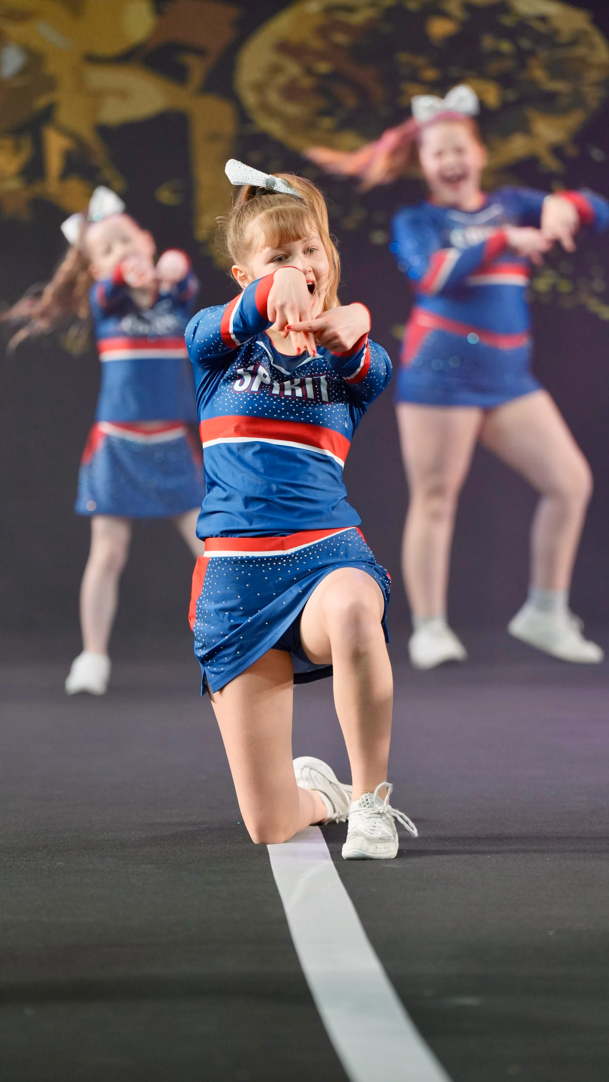 Young cheerleader kneeling on one knee, performing with two other cheerleaders blurred behind her.