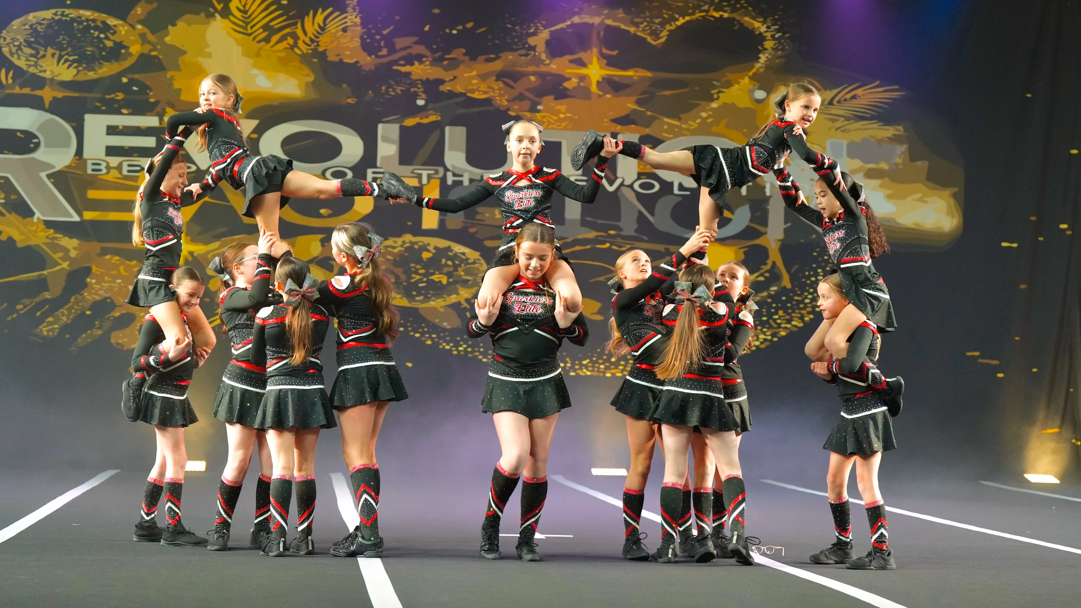Youth cheerleading team in black and red uniforms performing synchronized lifts on stage.