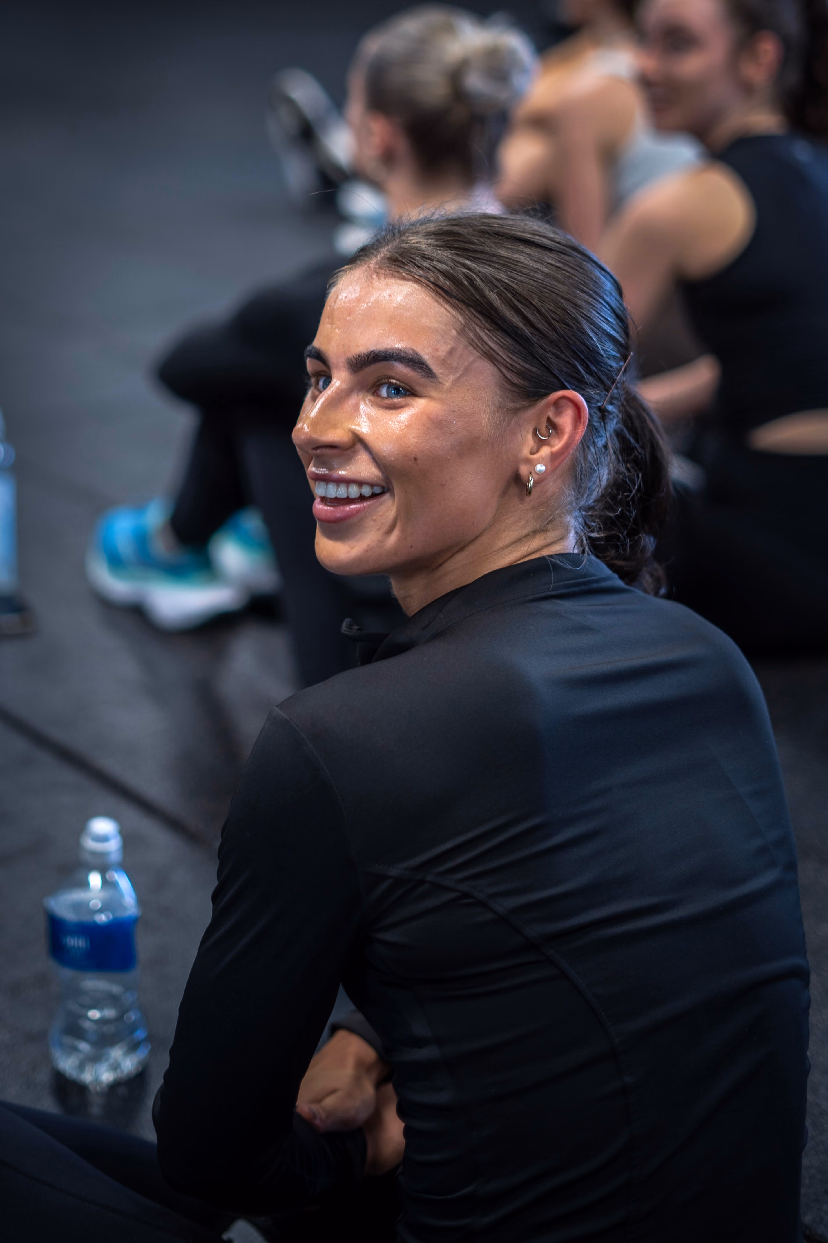 Smiling woman in black athletic wear sitting on gym floor with a water bottle nearby.