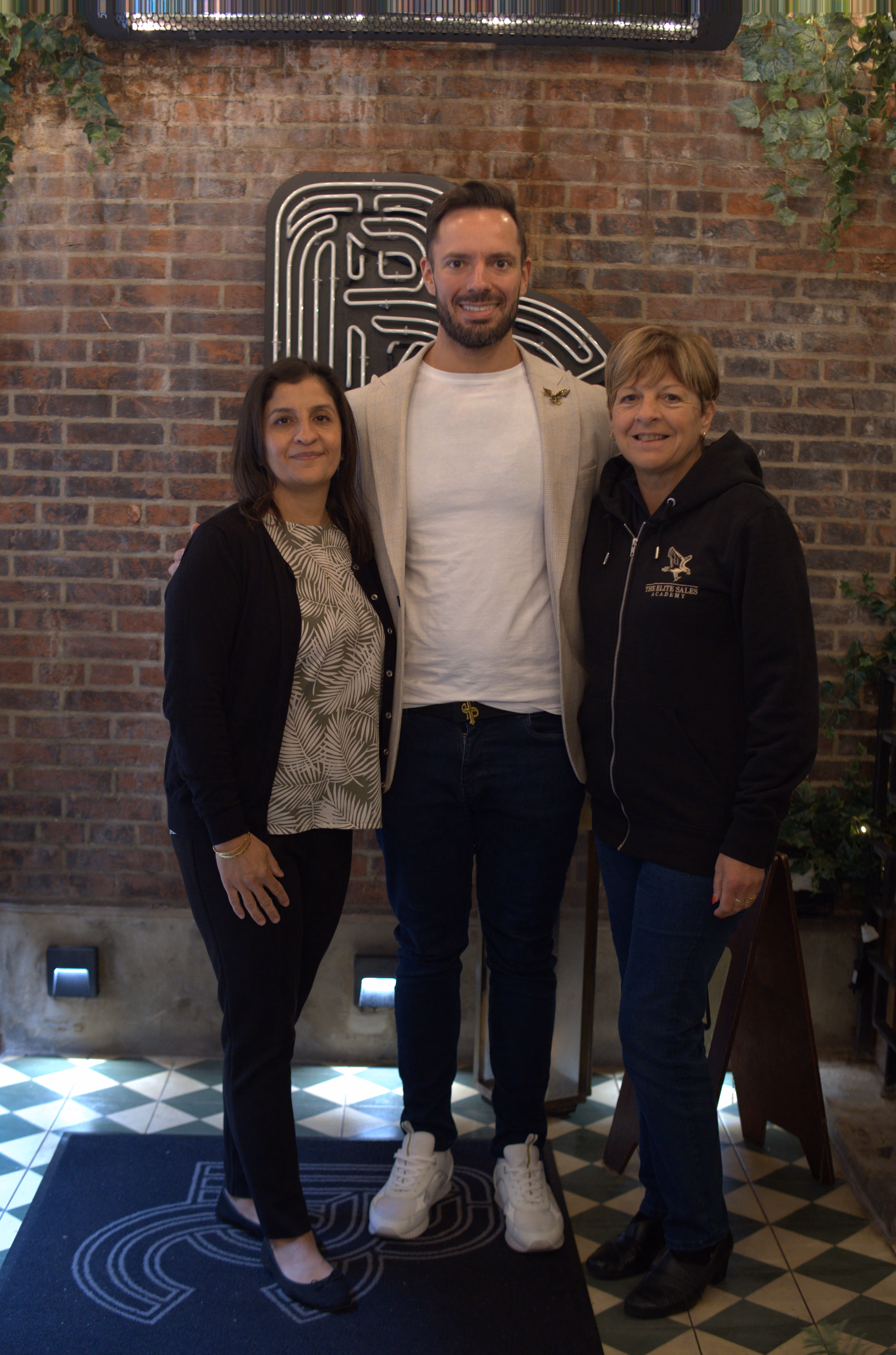 Three people standing together indoors against a brick wall and smiling at the camera.