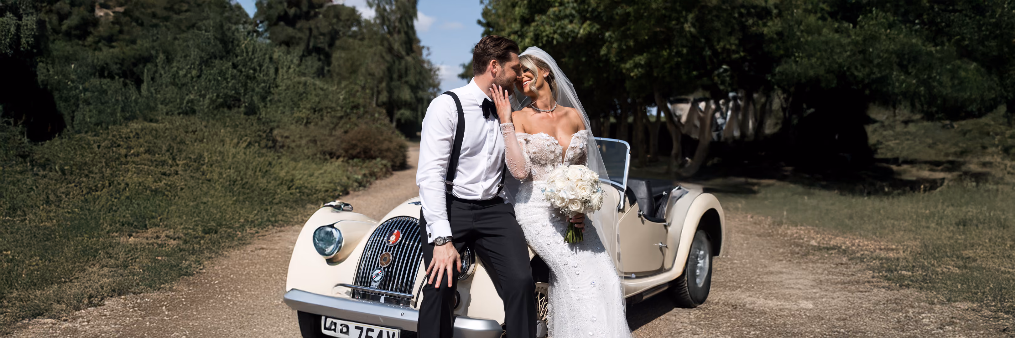 Bride in white dress and groom in suspenders share a kiss sitting on a vintage car outdoors.