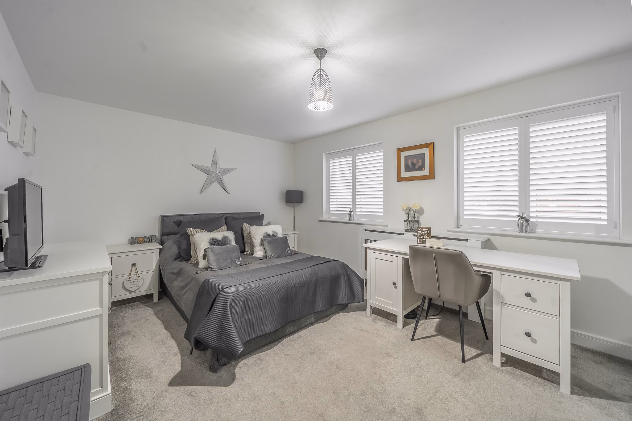 Modern bedroom with gray bedding, white furniture, wardrobe, desk, and two shuttered windows.