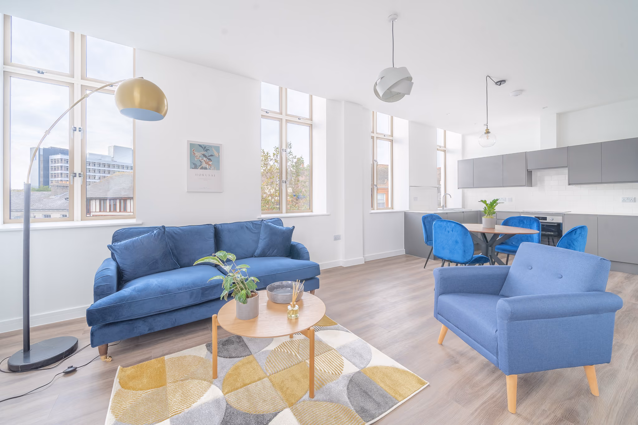 Bright living room with blue sofa, armchair, round table, geometric rug, and dining area with blue chairs.