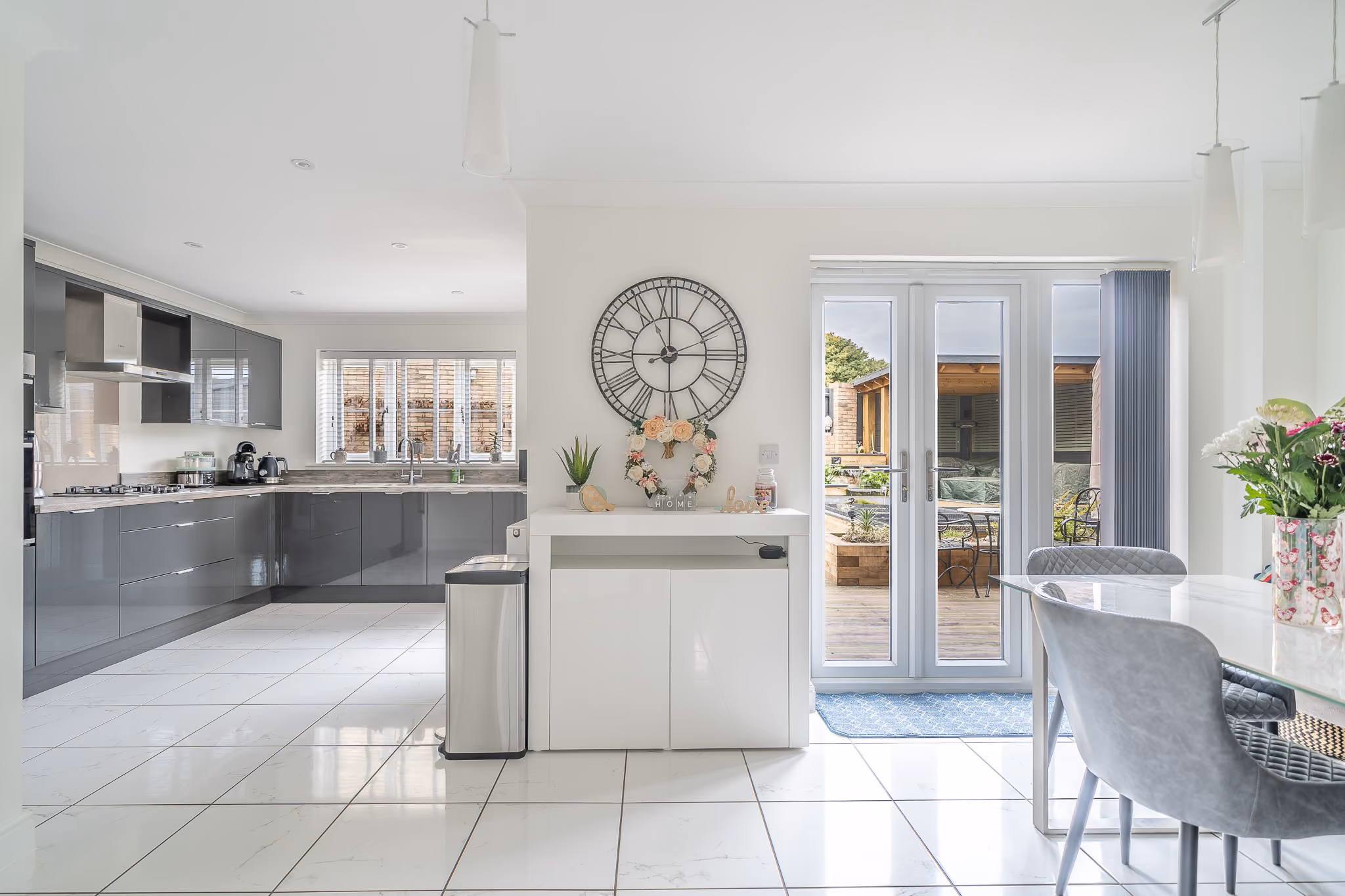 Bright modern kitchen with gray cabinets, large wall clock, glass doors, and a dining table with chairs.