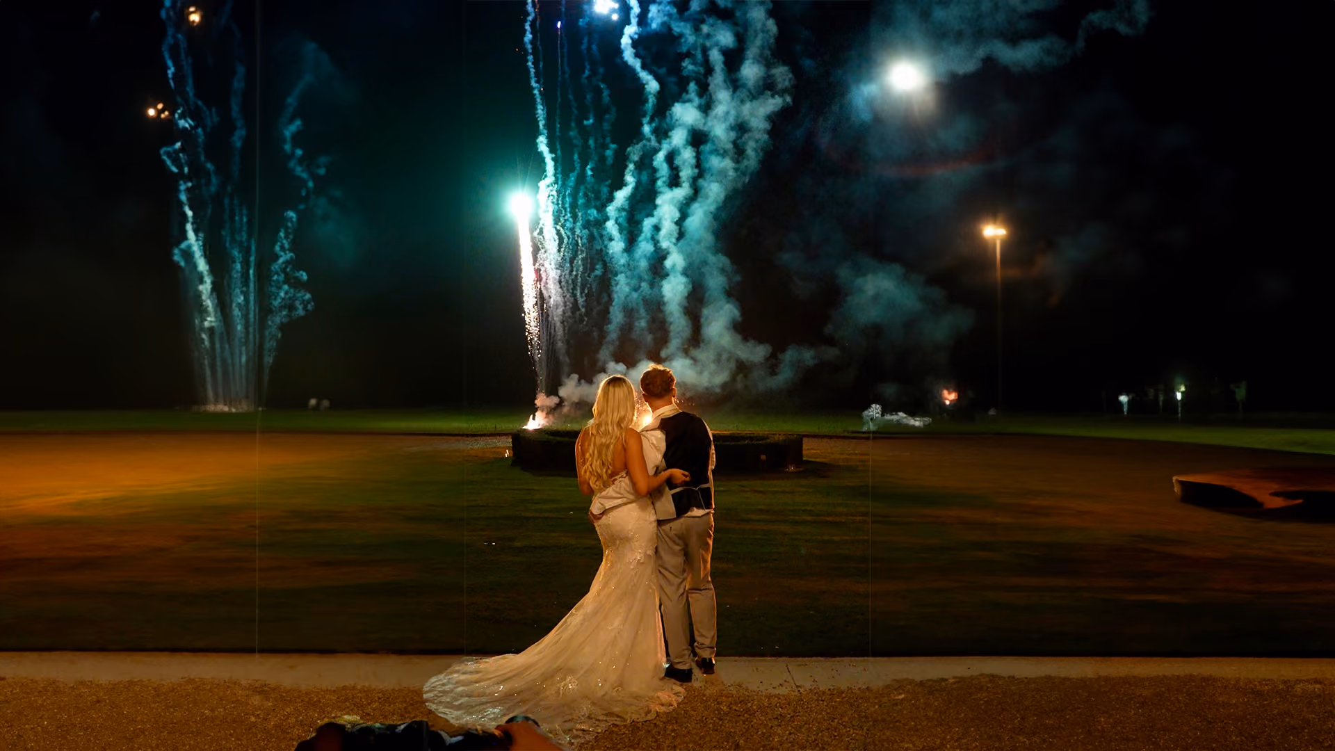 Couple in wedding attire standing closely, watching fireworks at night on a grassy field.