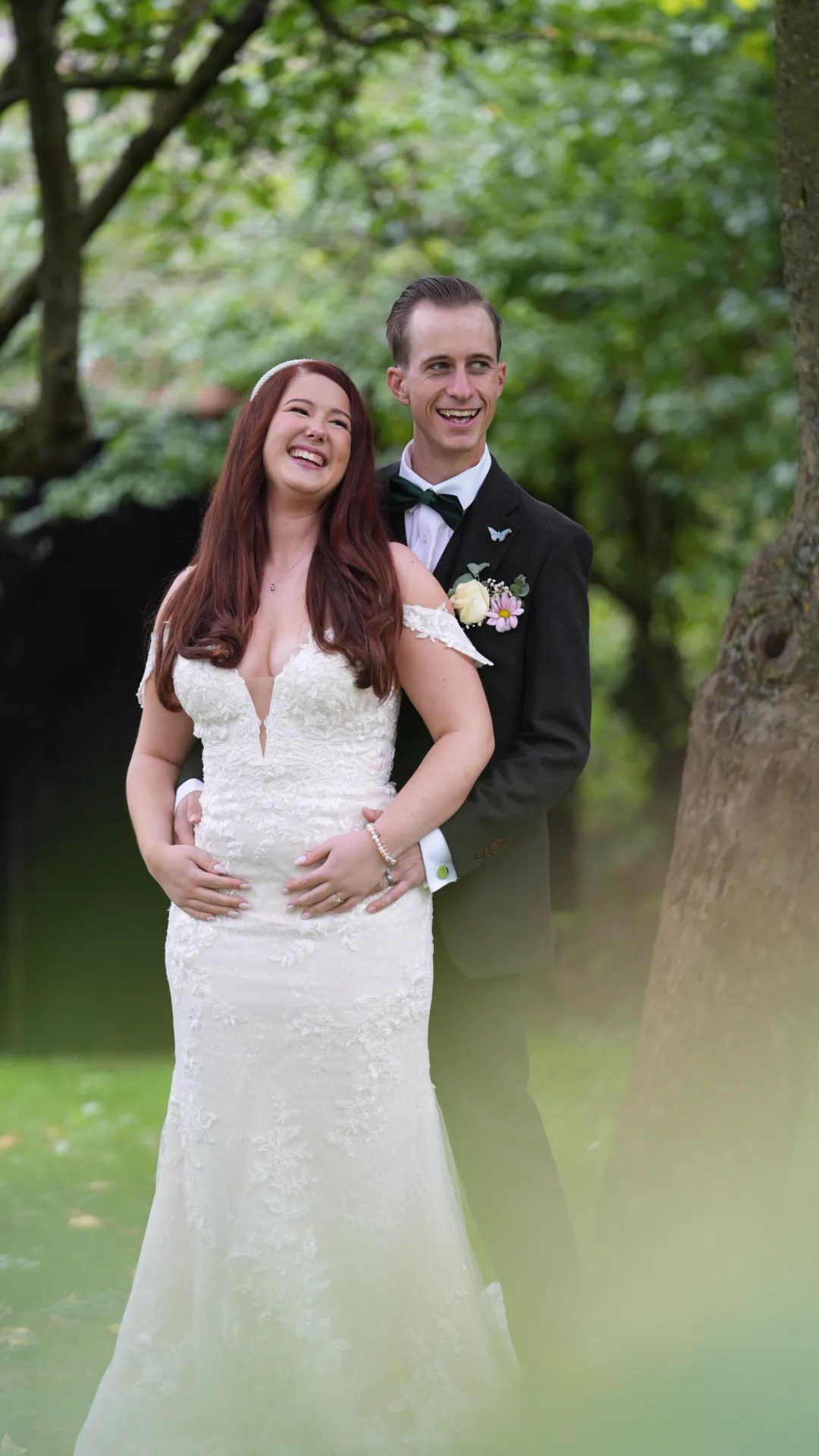 Bride and groom smiling outdoors, bride in lace dress, groom in dark suit with boutonniere.