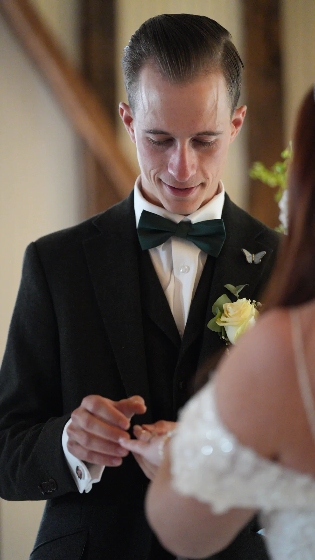 Groom in dark suit and bow tie placing a ring on bride's finger during wedding ceremony.