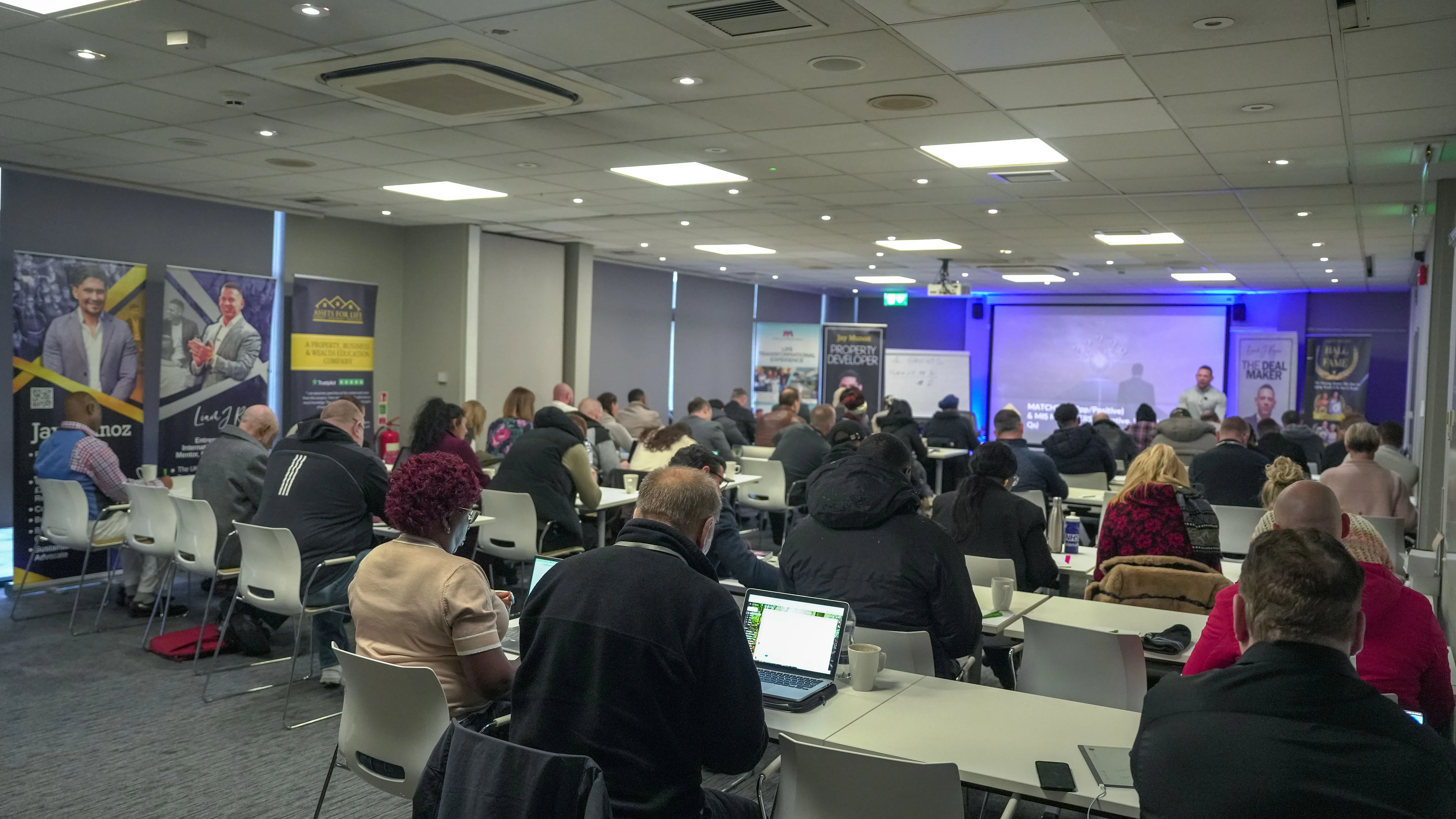 Audience of adults seated at desks listening to a speaker in a conference room with a projector screen.