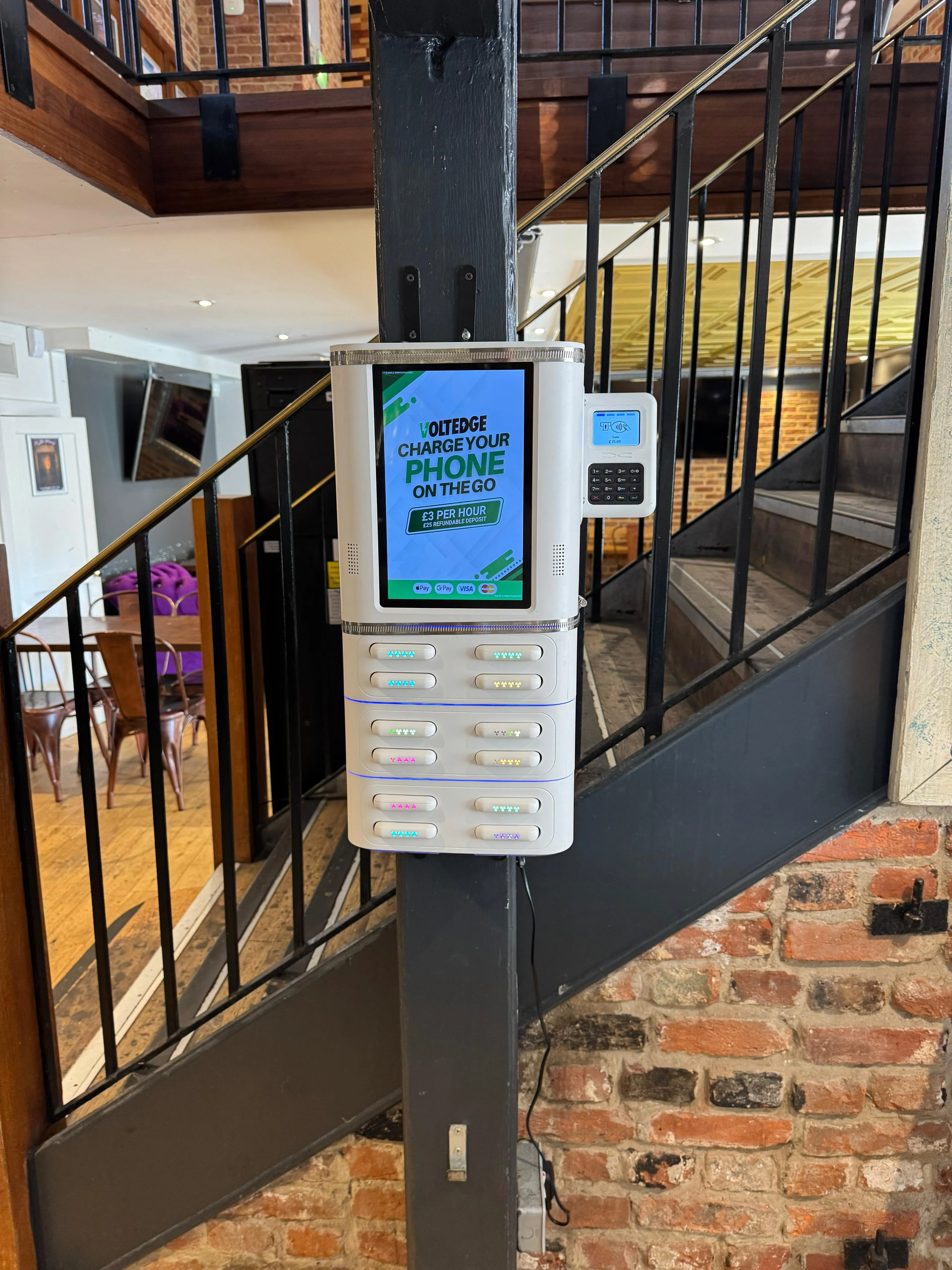 Wall-mounted phone charging station with lit lockers and payment keypad beside staircase and brick wall.