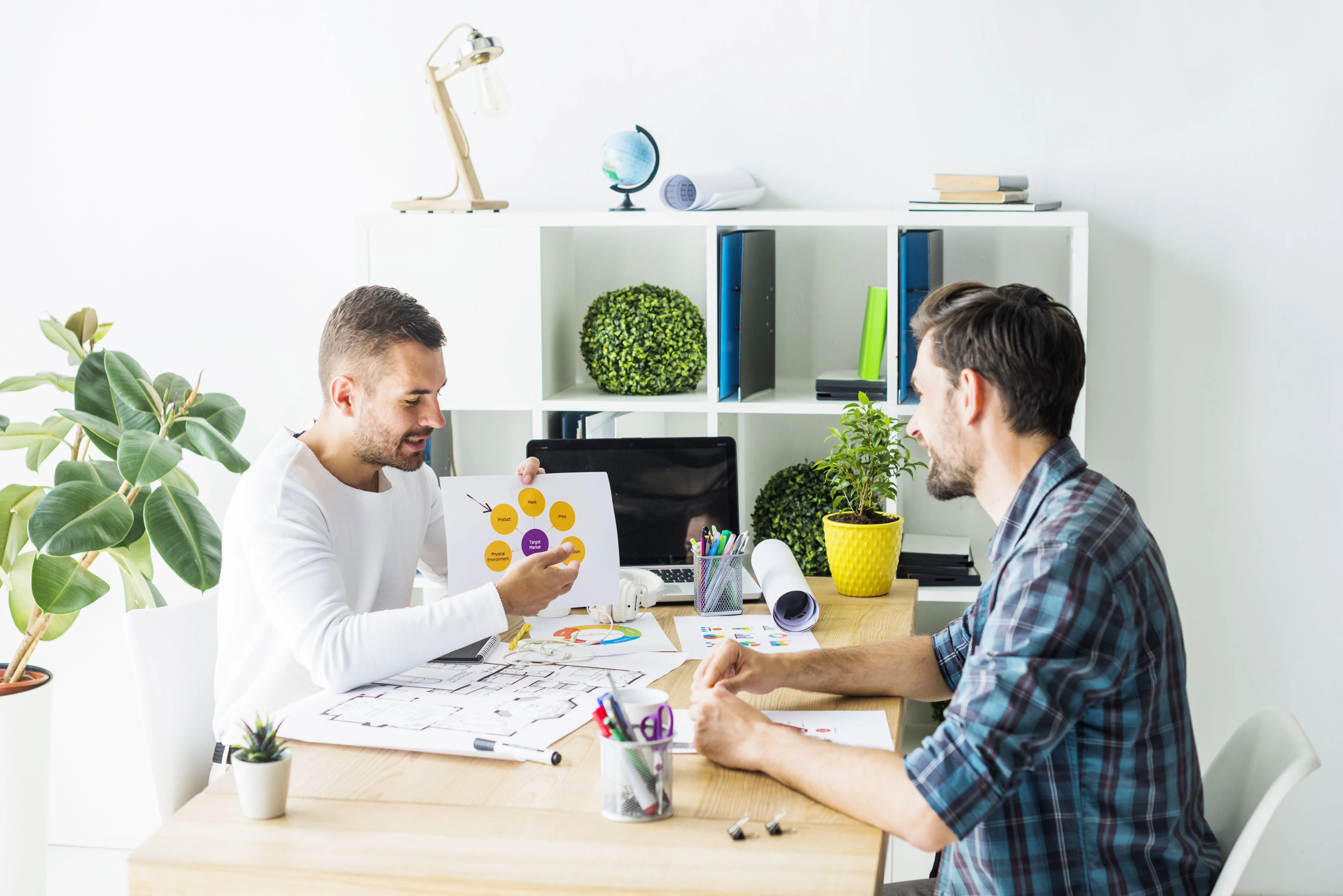 Two men at a desk, one showing a marketing diagram to the other in a bright office.