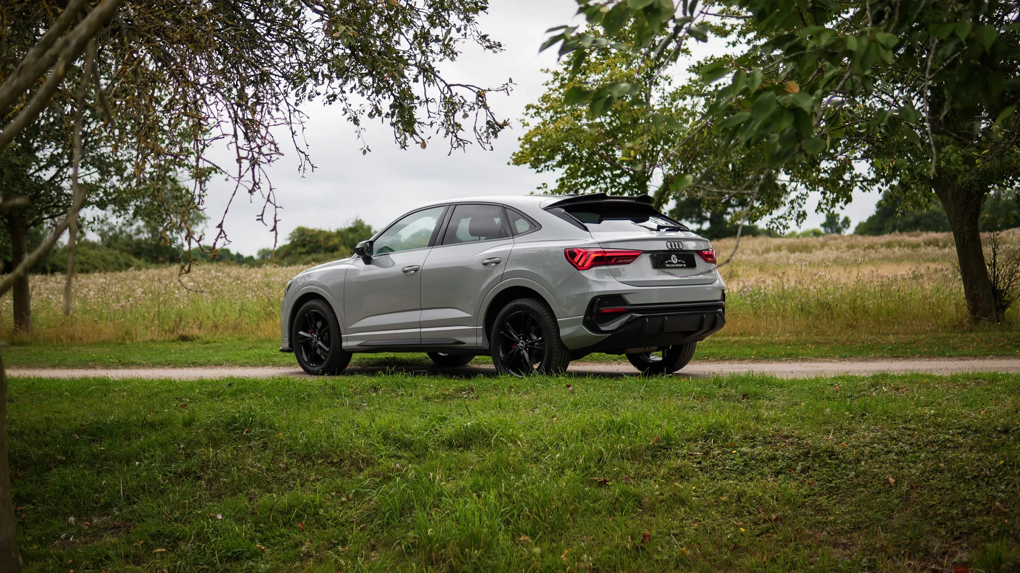 Silver Audi SUV parked on grass near a dirt path with trees and a field in the background.