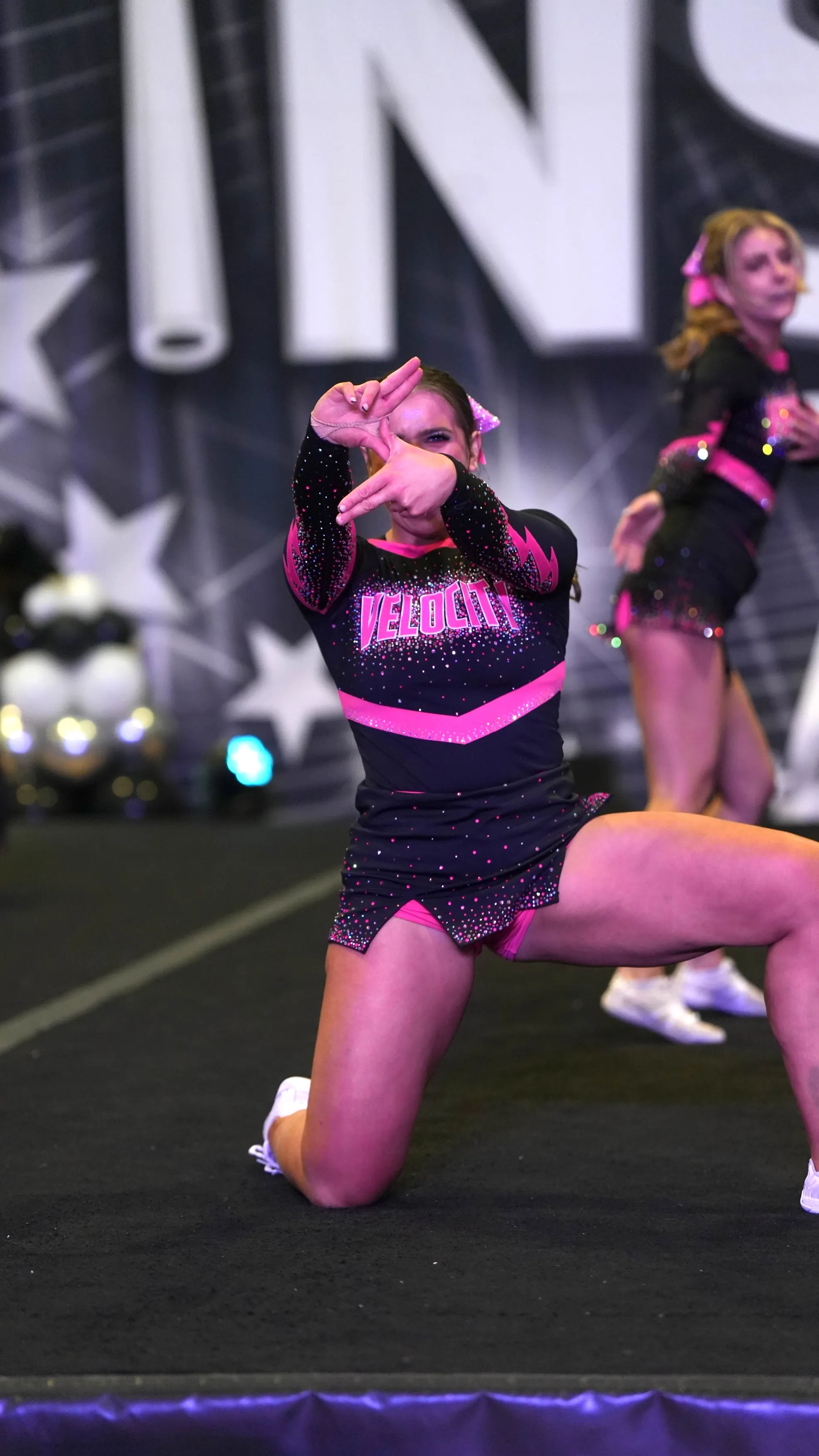 Female cheerleader in black and pink uniform kneeling and framing face with hands during performance.