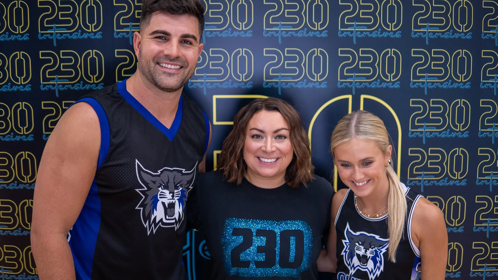 Smiling man and two women wearing sports jerseys and a 230 intensives shirt in front of a 230 intensives backdrop.