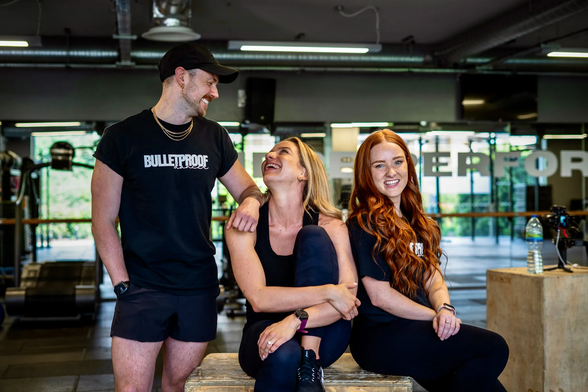 Three smiling people in athletic wear in a gym, two women sitting on a wooden box and one man standing.