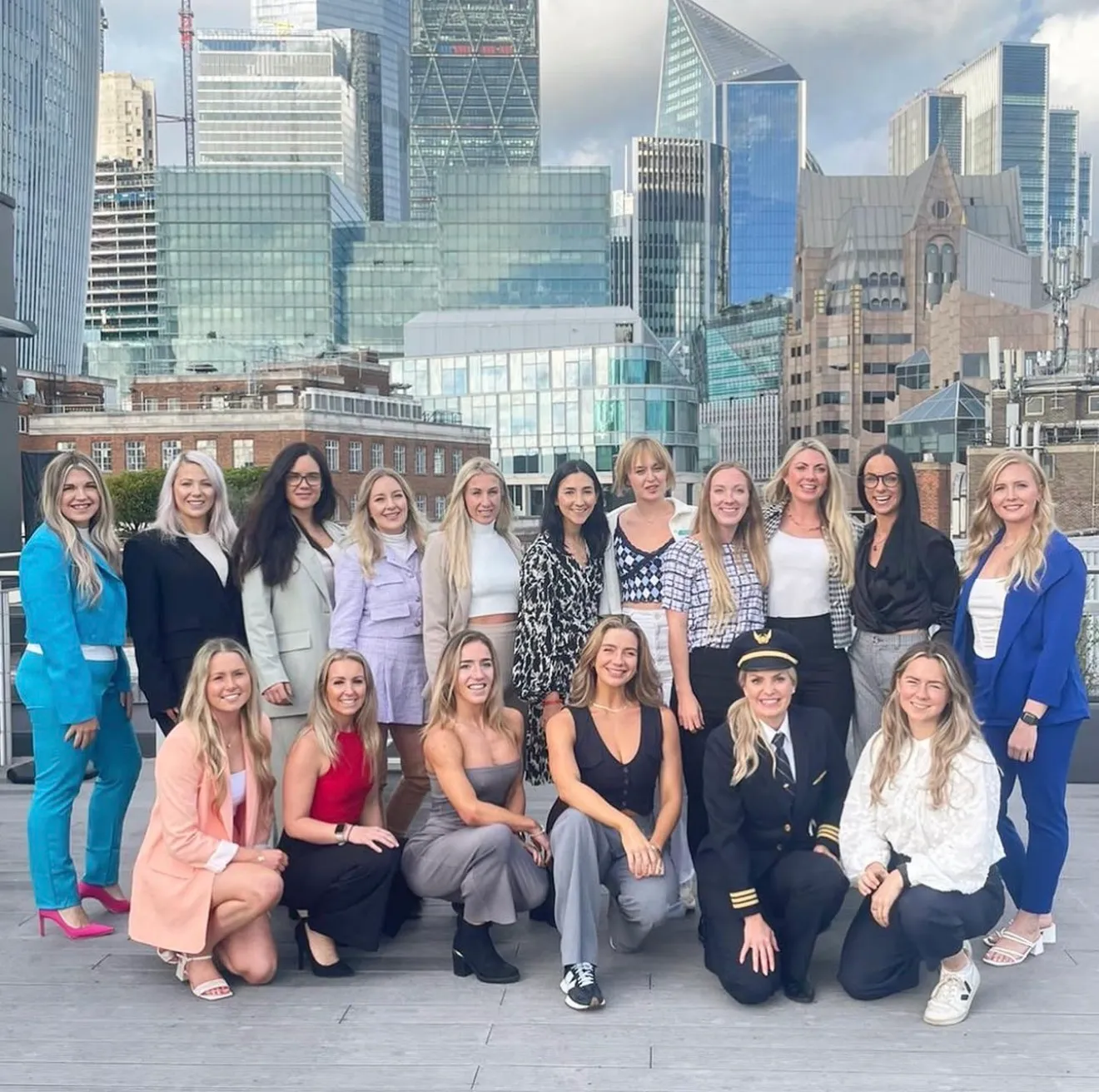 Group of 18 women posing outdoors with a city skyline of modern buildings in the background.