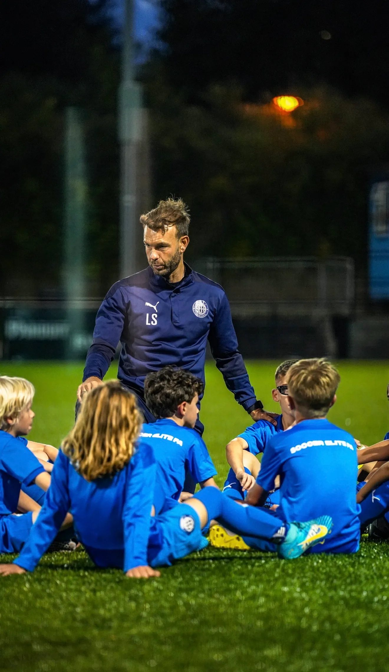 Coach giving instructions to young soccer players sitting on grass field at night.