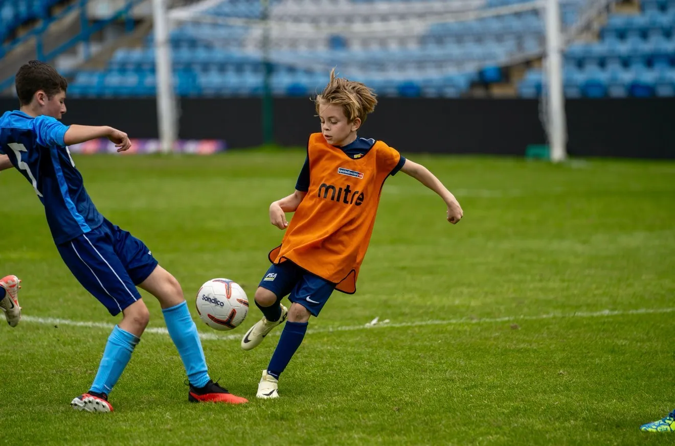Two boys playing soccer on a field, one in blue and the other in an orange vest kicking the ball.