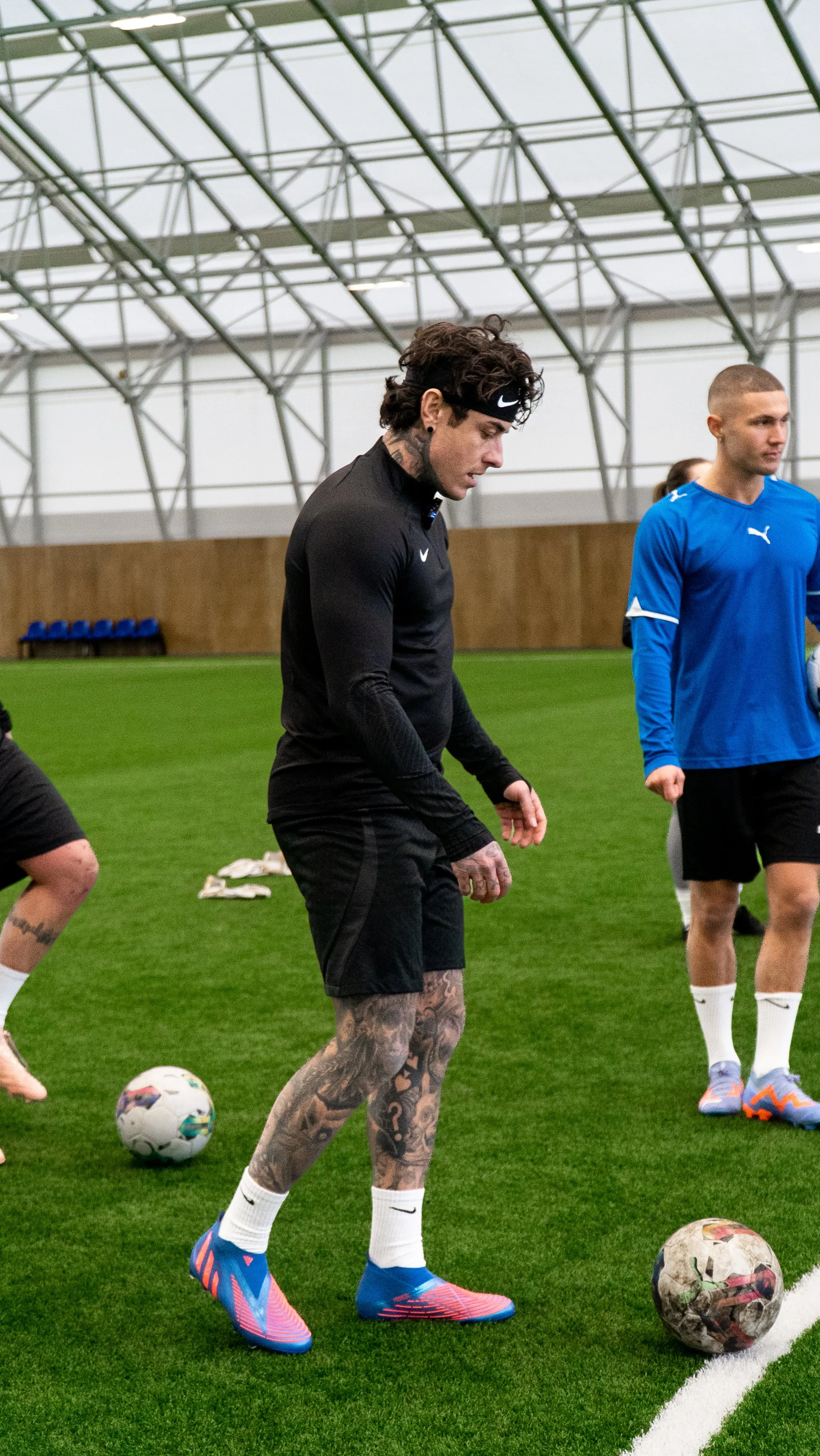 Tattooed man in black sportswear and blue cleats prepares to kick a soccer ball on an indoor field.
