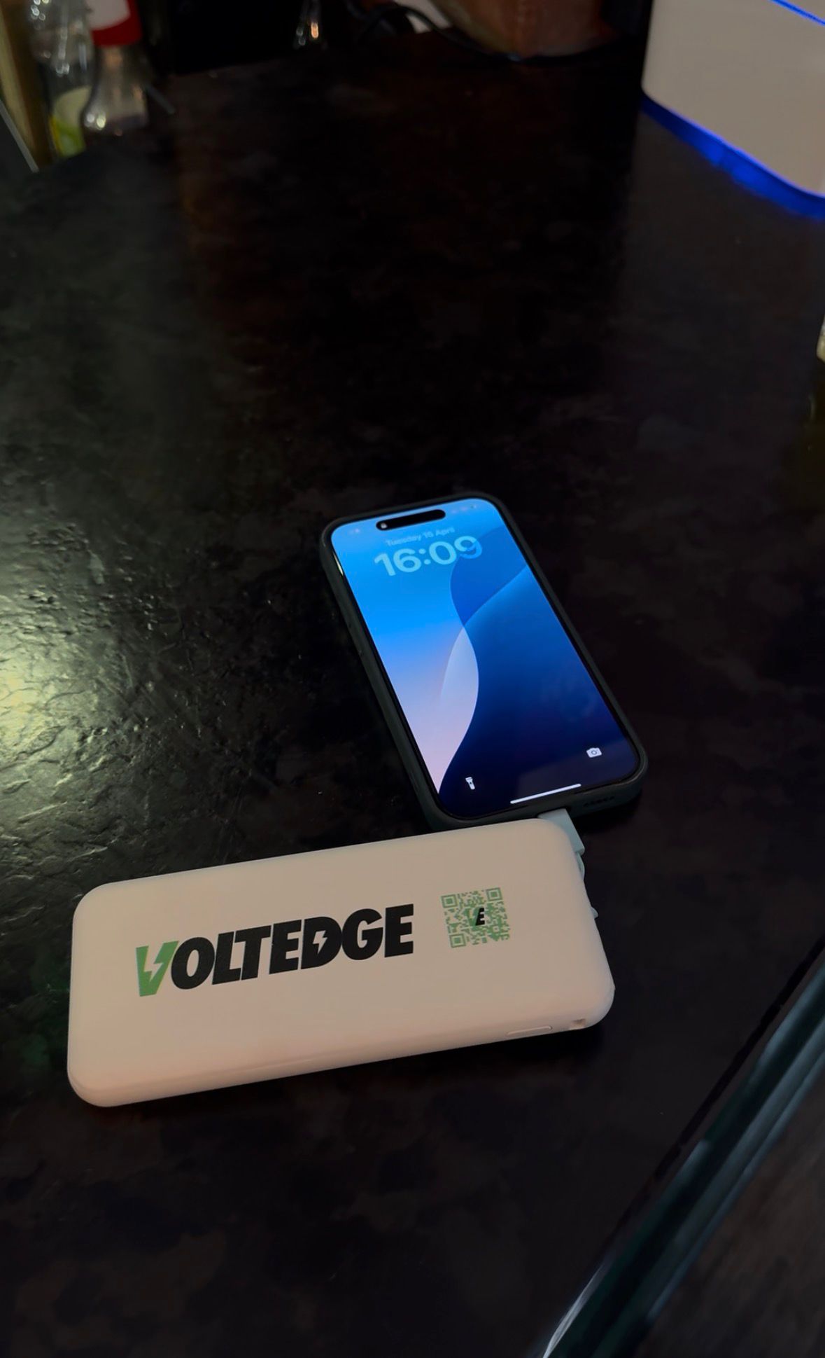 An iPhone with a blue abstract wallpaper is charging from a white power bank labeled VOLTEDGE on a dark surface.