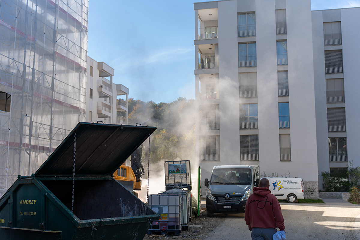 Man in a red hoodie walking near construction site with trucks, containers, and smoke in front of modern apartment buildings.