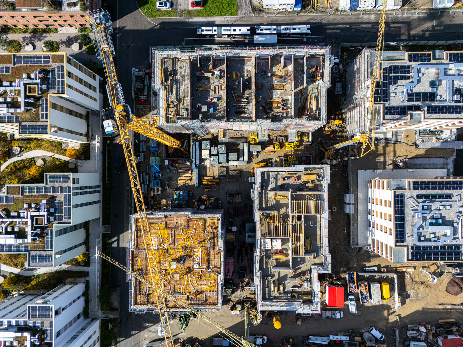 Aerial view of a construction site with cranes, building foundations, construction materials, and surrounding completed buildings with solar panels.