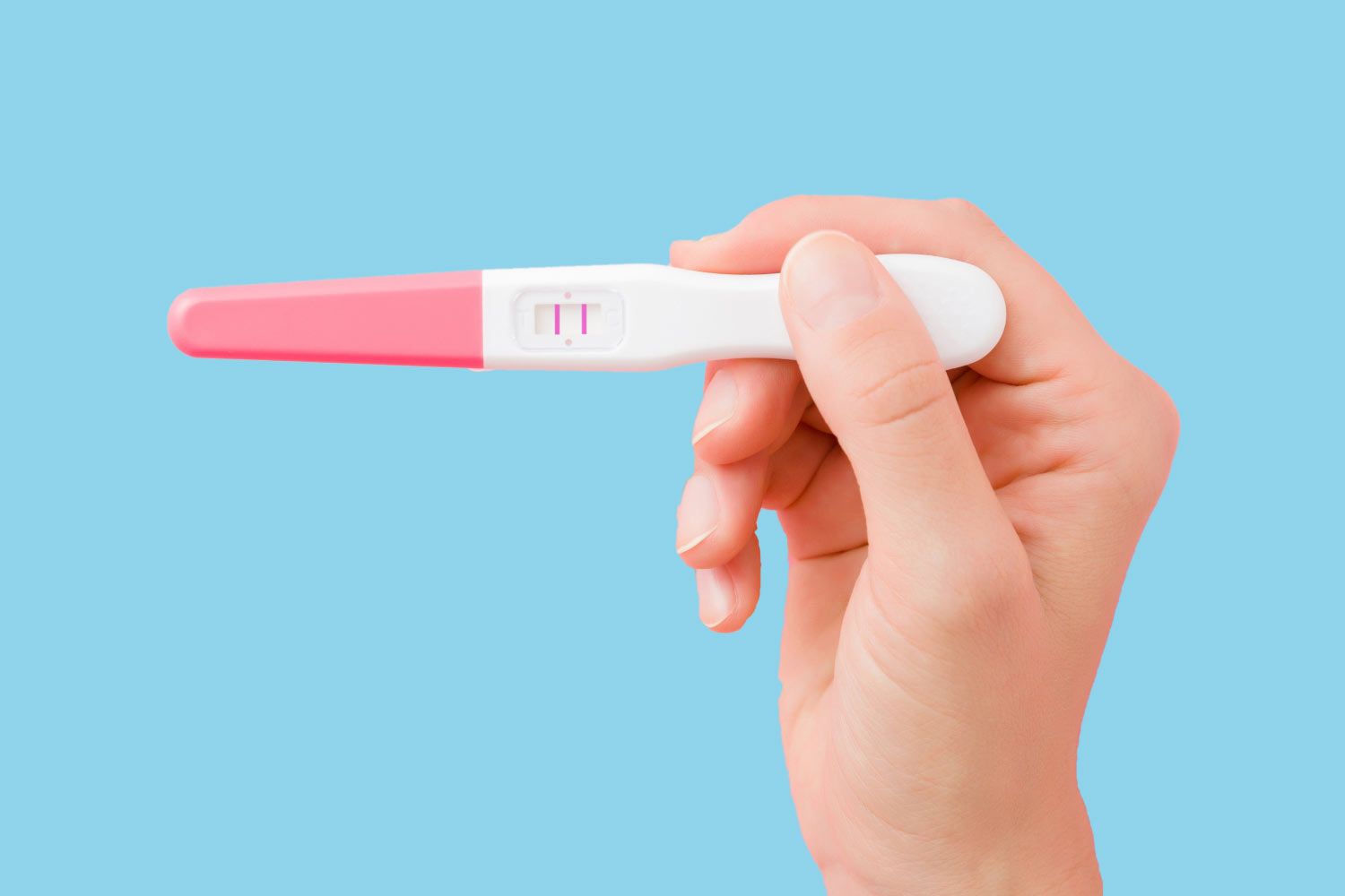 Hand holding a positive pregnancy test showing two pink lines against a light blue background.