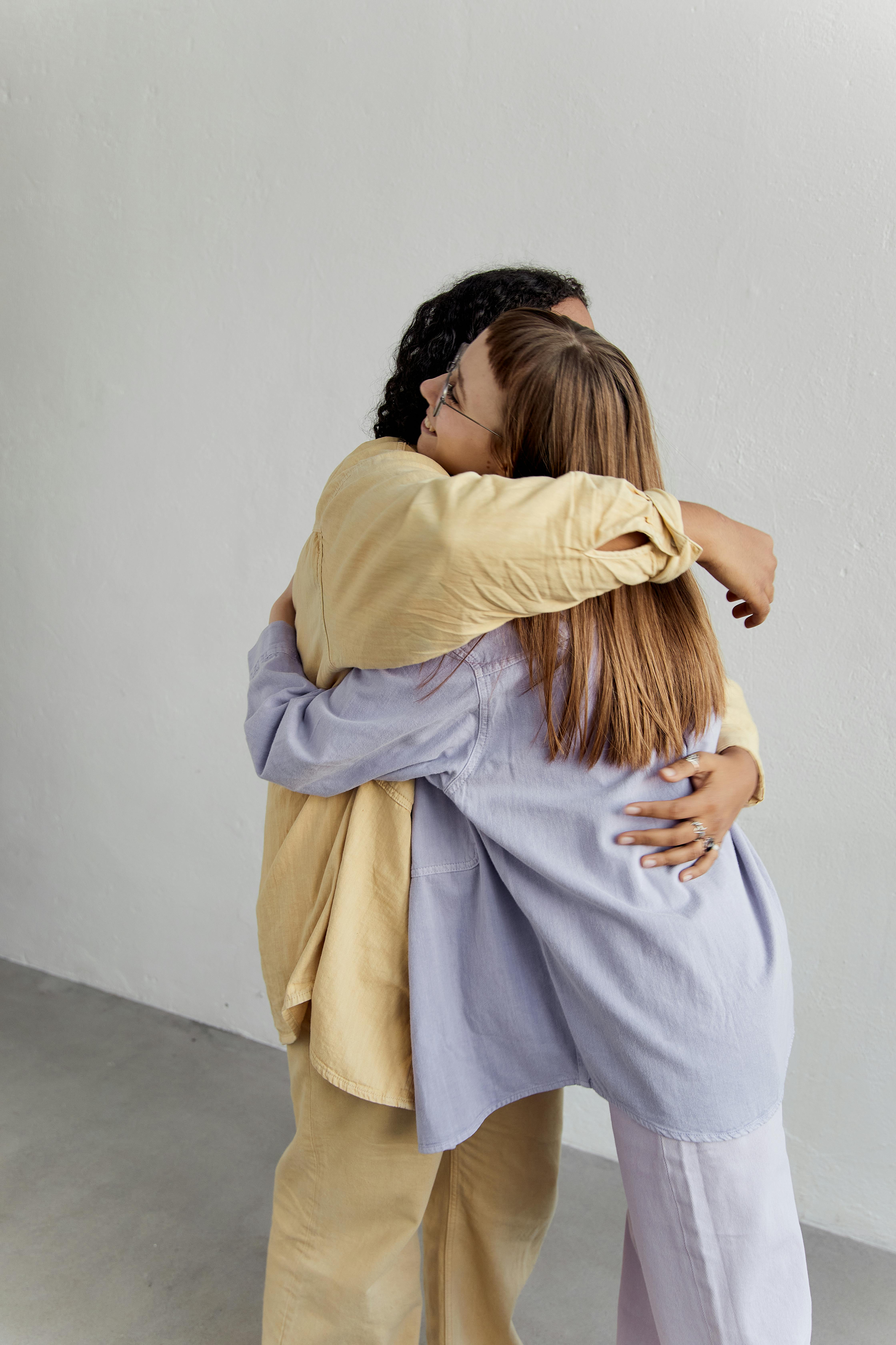 Two women hugging each other closely with smiles, one wearing a yellow outfit and the other in a light purple outfit.