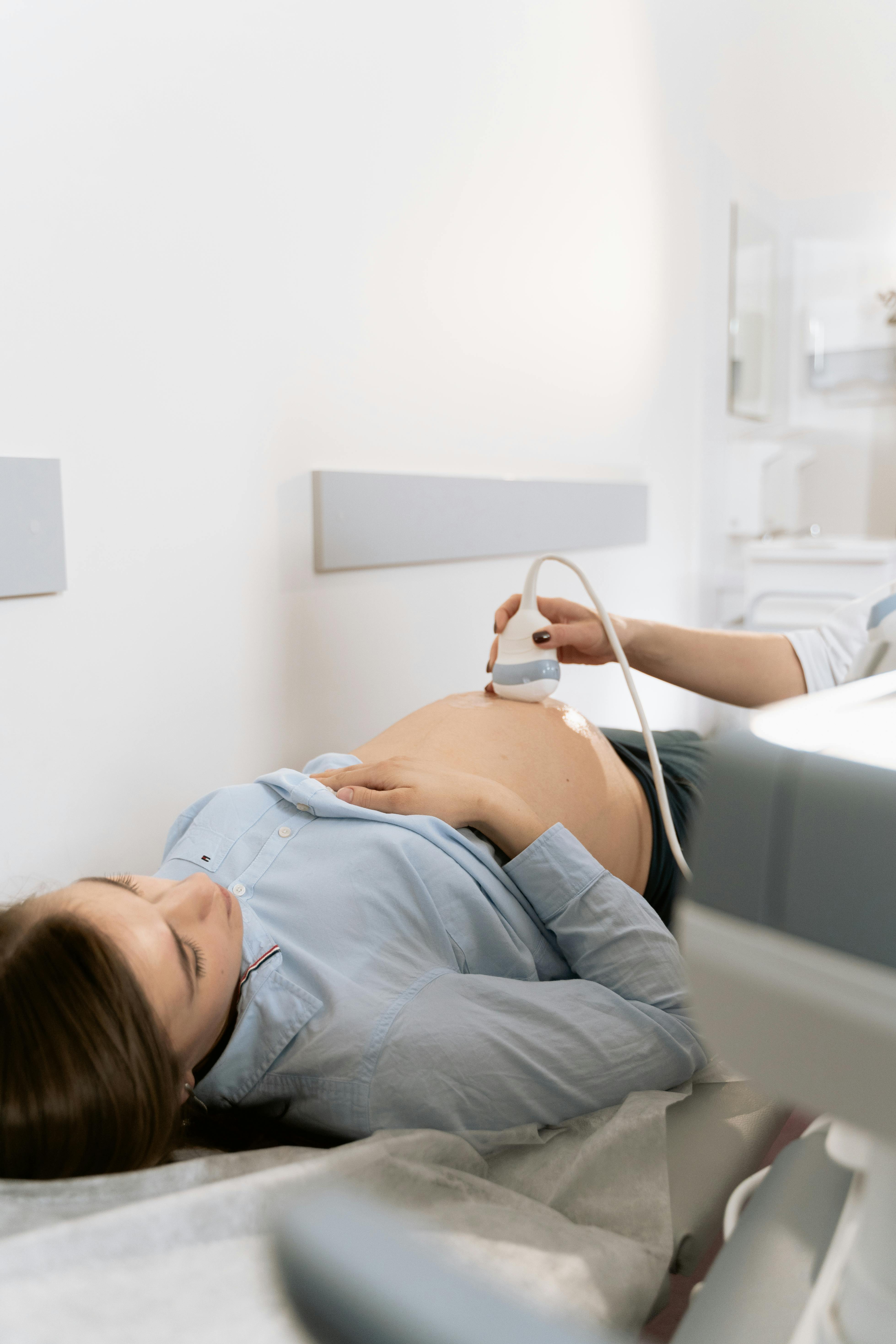 Pregnant woman lying on a medical bed receiving an ultrasound scan on her bare belly in a clinic.