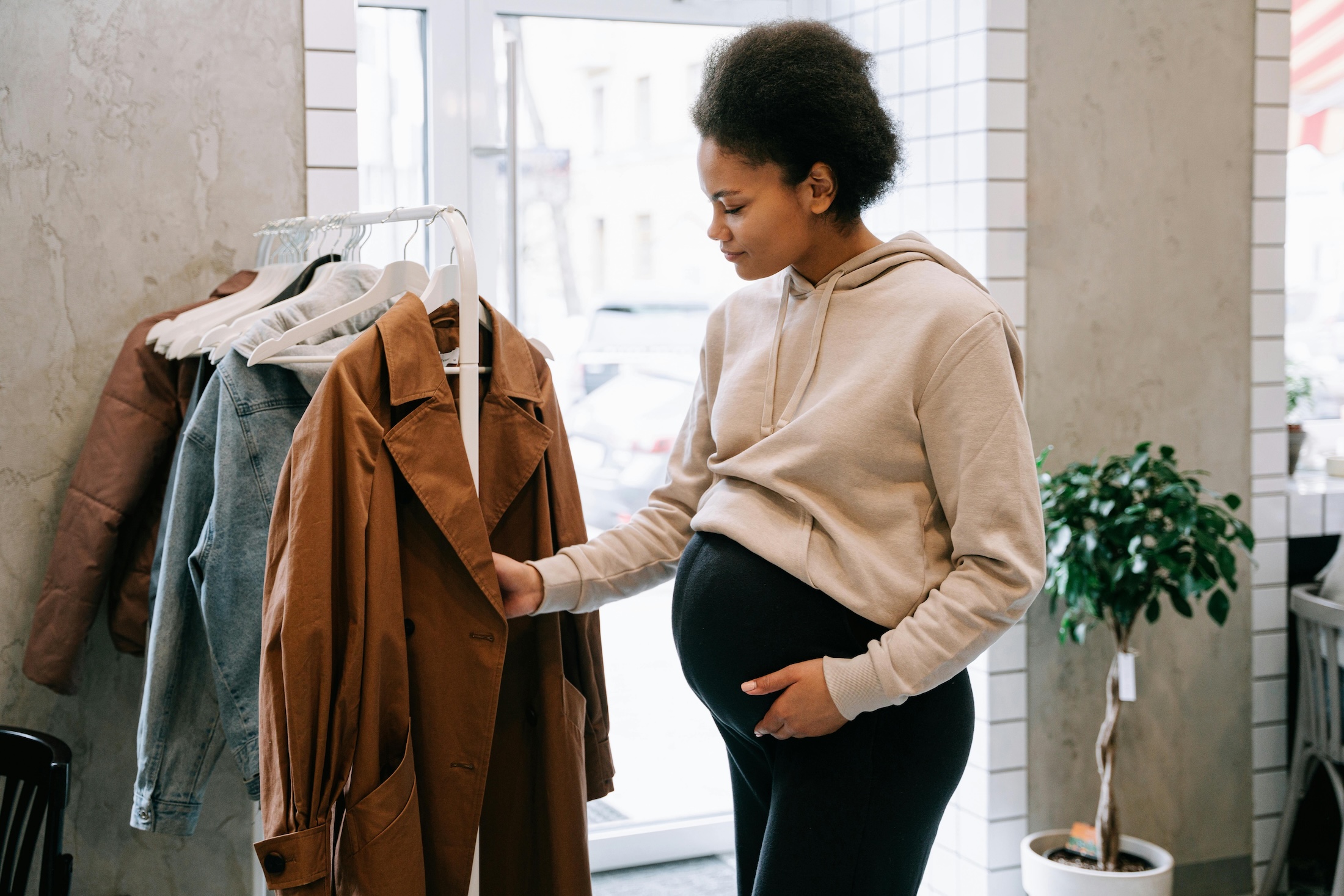 Pregnant woman wearing a beige hoodie touching a brown coat on a clothing rack in a store.
