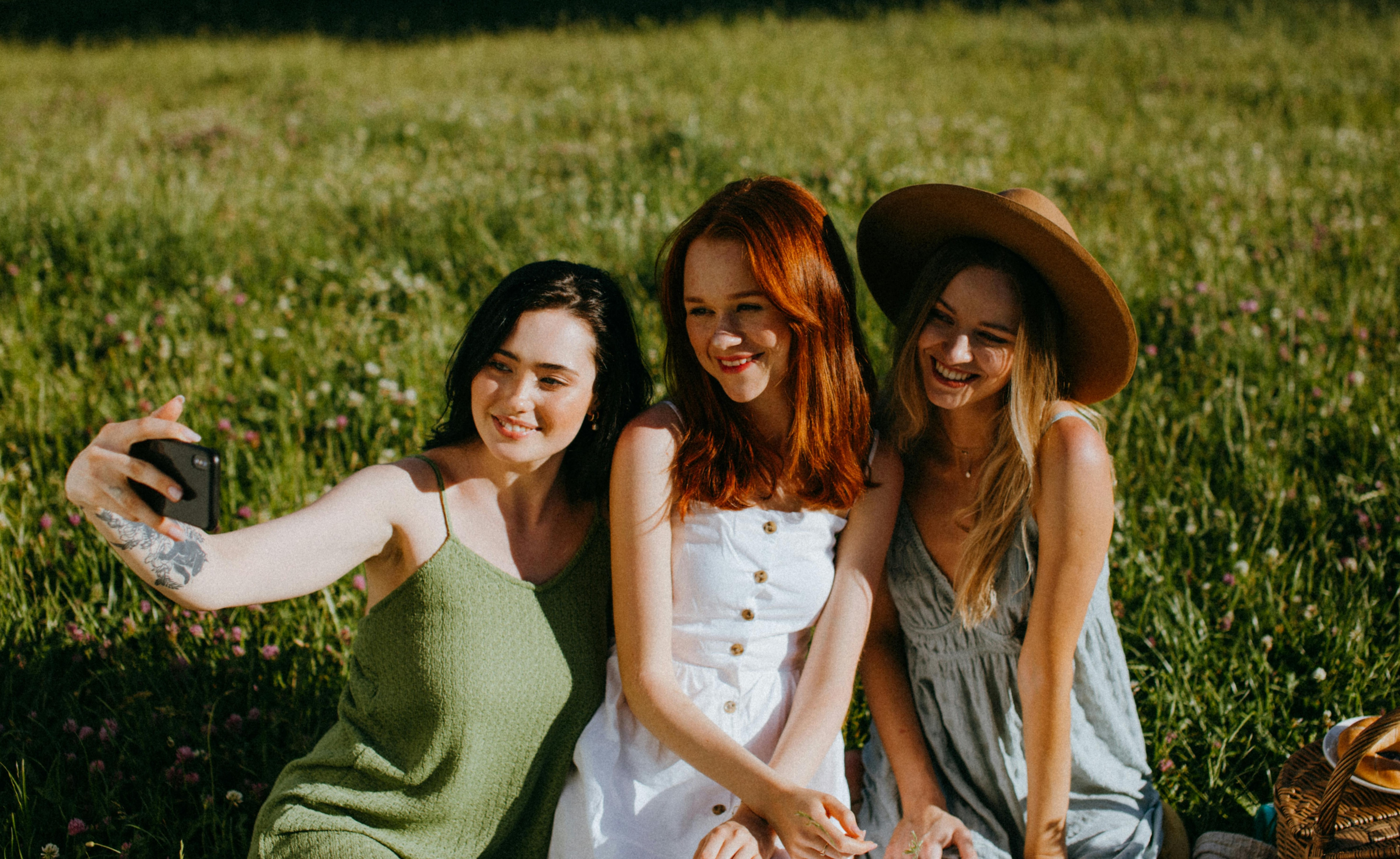 Three young women sitting in a sunny field taking a selfie, one wearing a green dress, one in a white dress, and one in a gray dress with a brown hat.