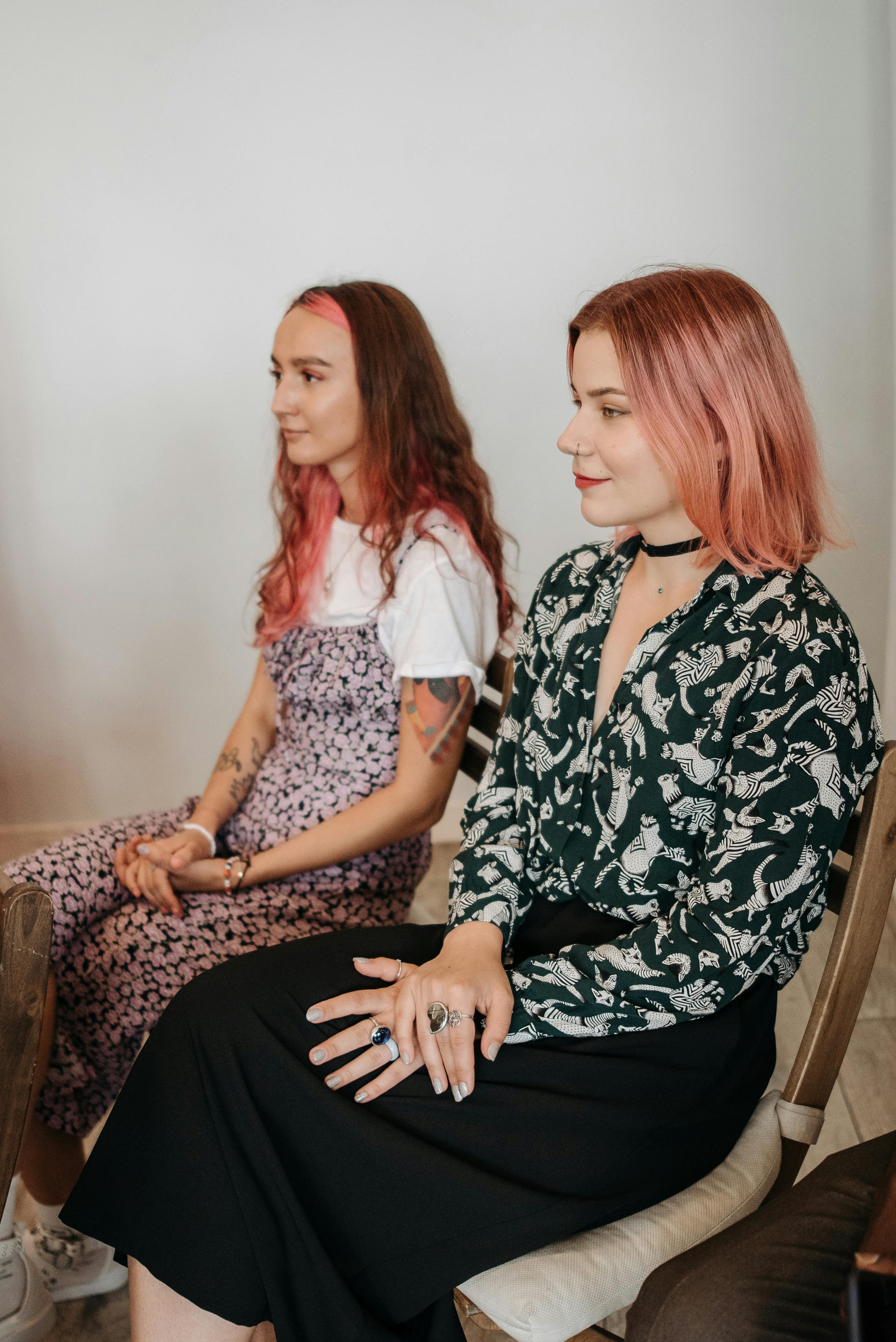 Two young women with pink hair sitting side by side on wooden chairs against a plain light background.