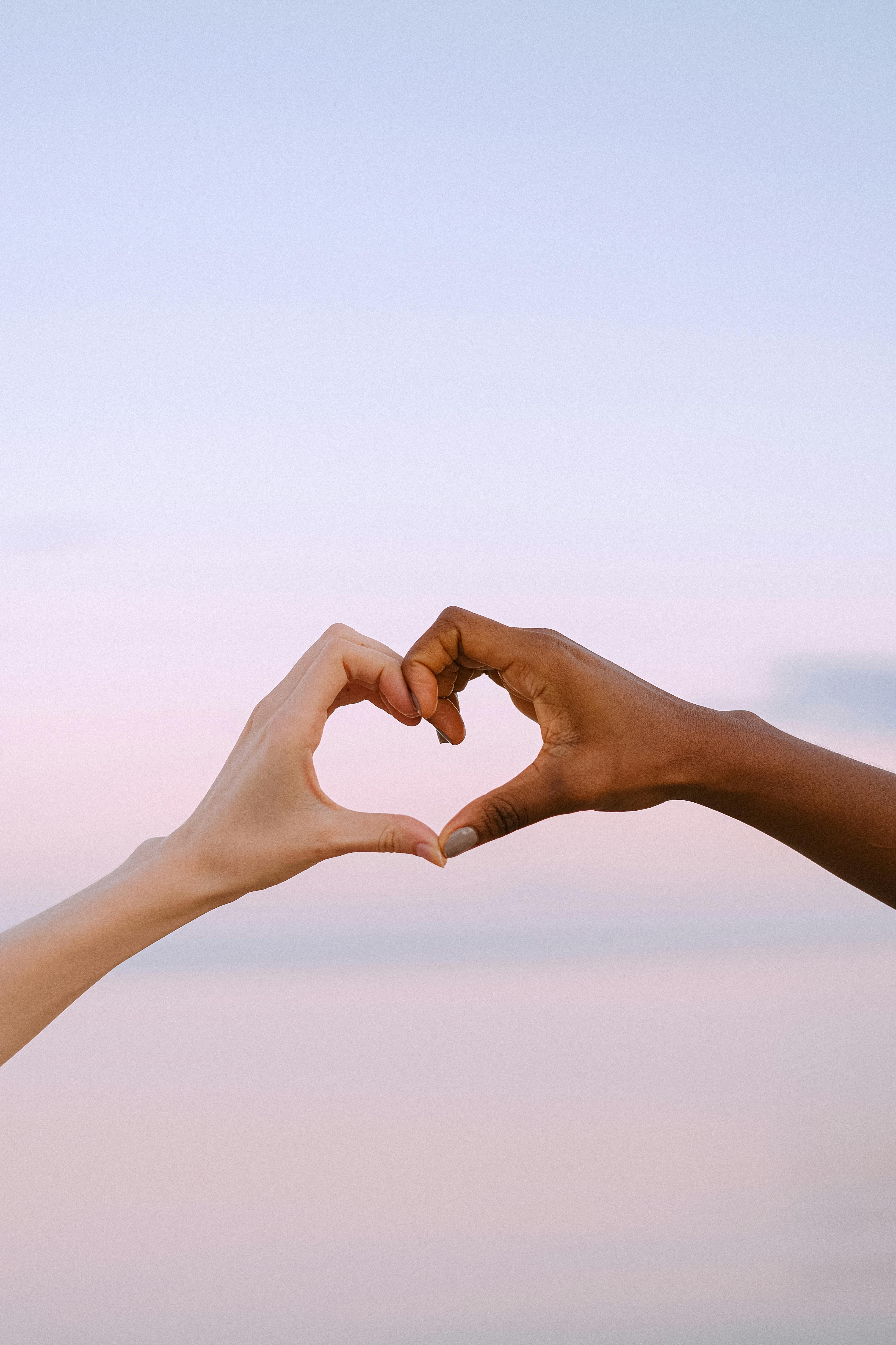 Two hands of different skin tones forming a heart shape against a pastel sky background.