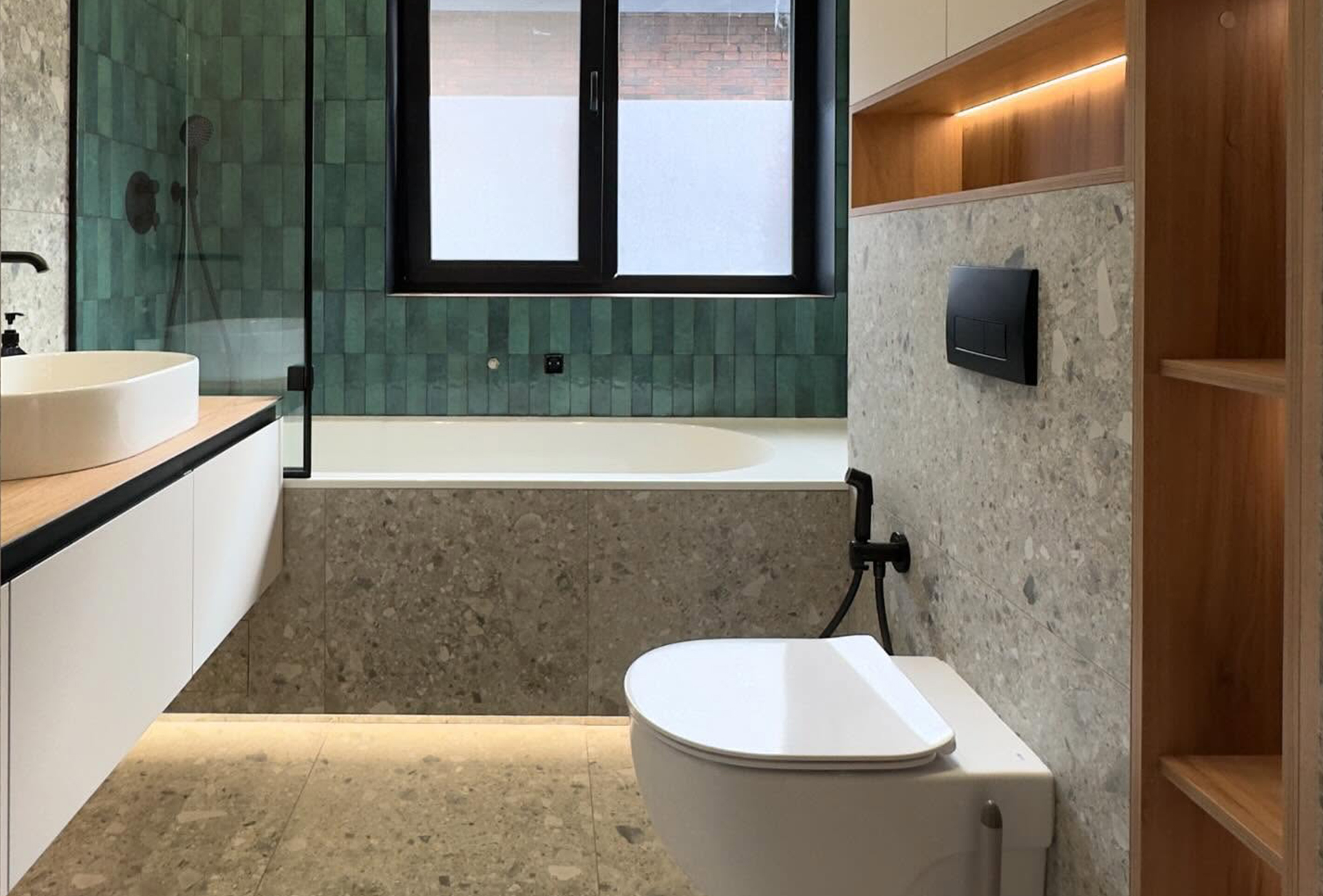 Сучасний дизайн ванної кімнати з підвісним унітазом, підсвіченою тумбою під раковину та ванною, облицьованою сірим терраццо, акцентна стіна над ванною з зеленої глянцевої плитки "кабанчик".