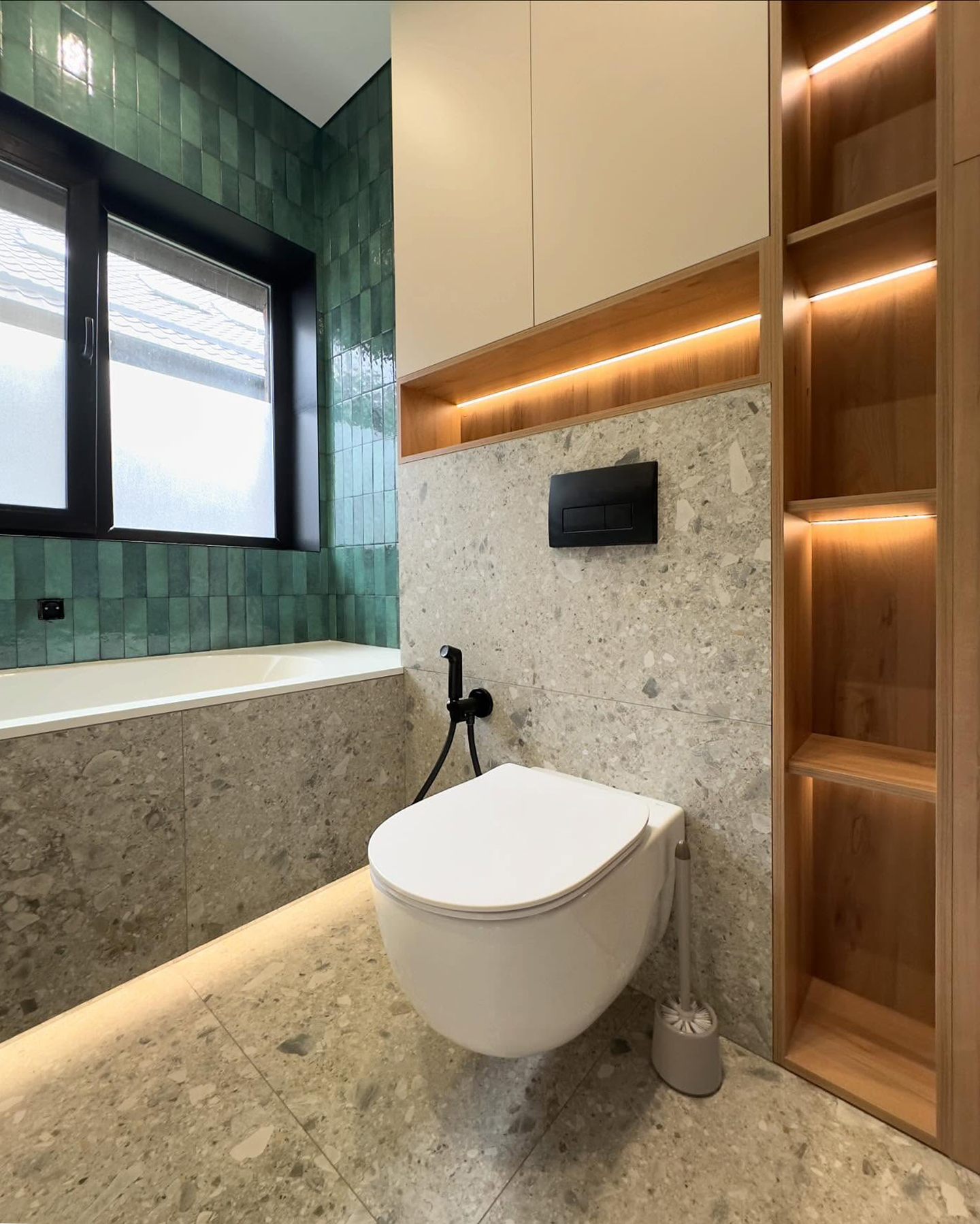 Сучасна ванна кімната: підвісний унітаз, вбудована шафа з білими глянцевими фасадами та відкритими дерев'яними полицями з підсвічуванням, стіни облицьовані сірою плиткою під терраццо.