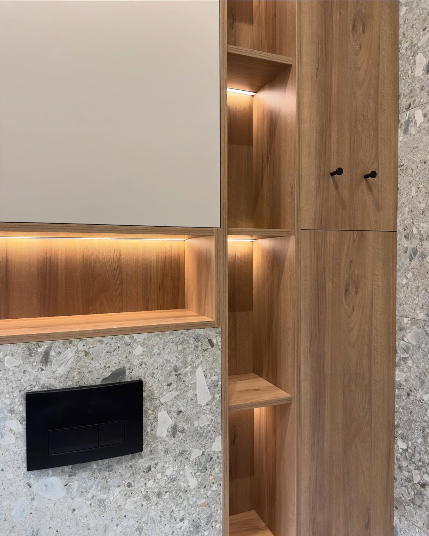 Вертикальний фрагмент вбудованих меблів: білі закриті шафи, відкриті дерев'яні полиці з підсвічуванням, шафа з дерев'яними фасадами та чорними мінімалістичними ручками на тлі сірої стіни під терраццо.