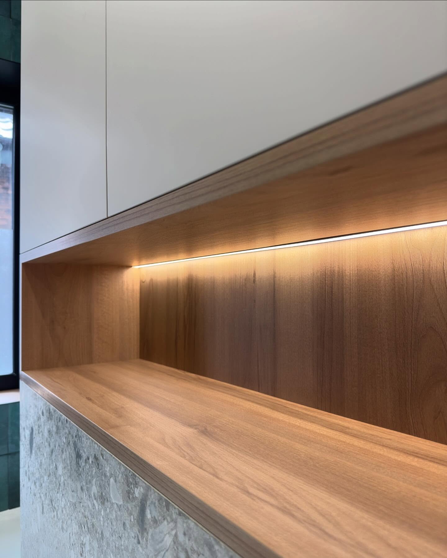Крупний план дерев'яної ніші з інтегрованим LED-підсвічуванням над зоною унітазу, частина вбудованої шафи з білим фасадом та стіна з сірої плитки під терраццо.