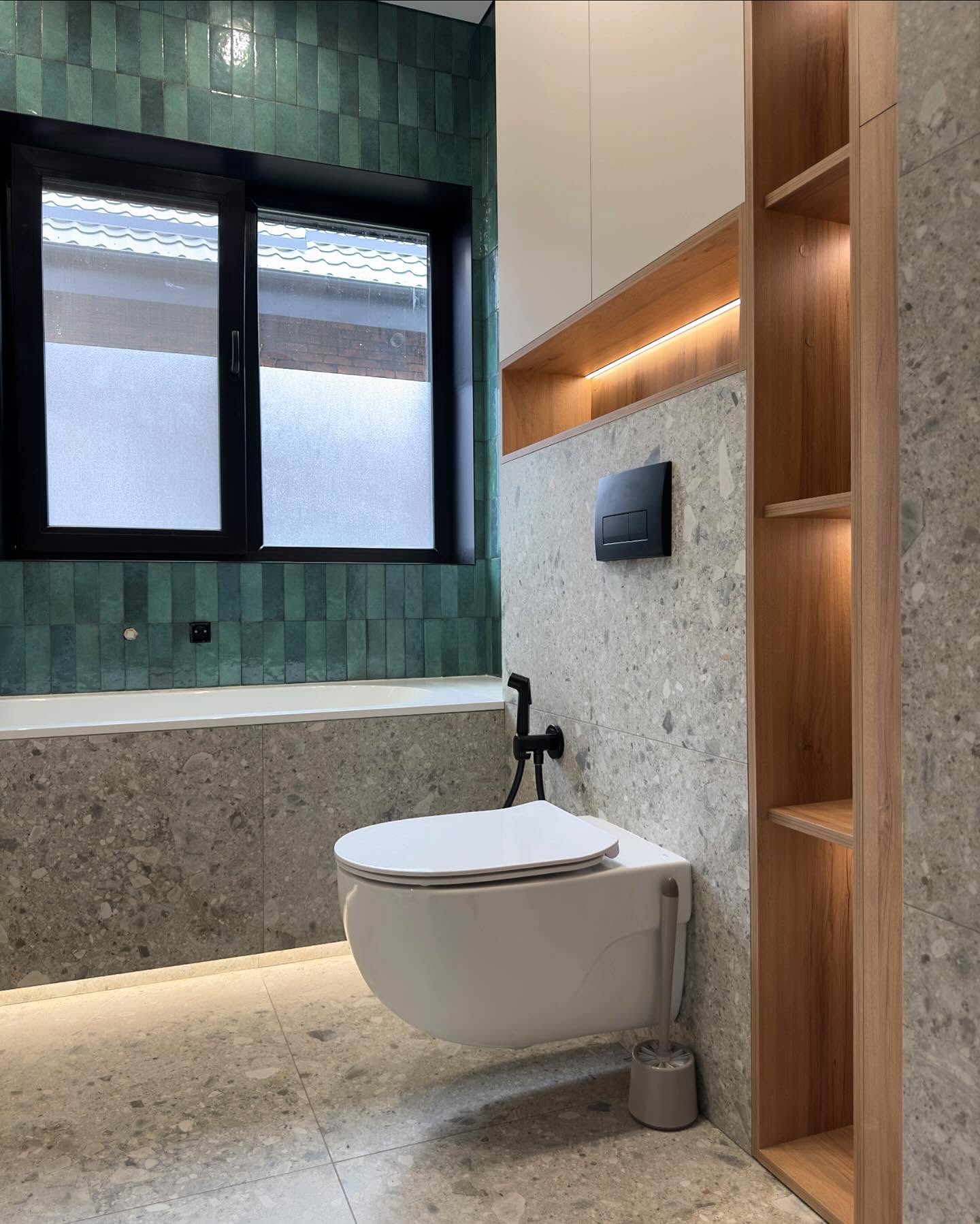 Підвісний унітаз з чорною панеллю змиву та гігієнічним душем, вбудовані дерев'яні полиці з підсвічуванням, сіра плитка під терраццо на підлозі та стіні, вид на вікно та зелену плитку.