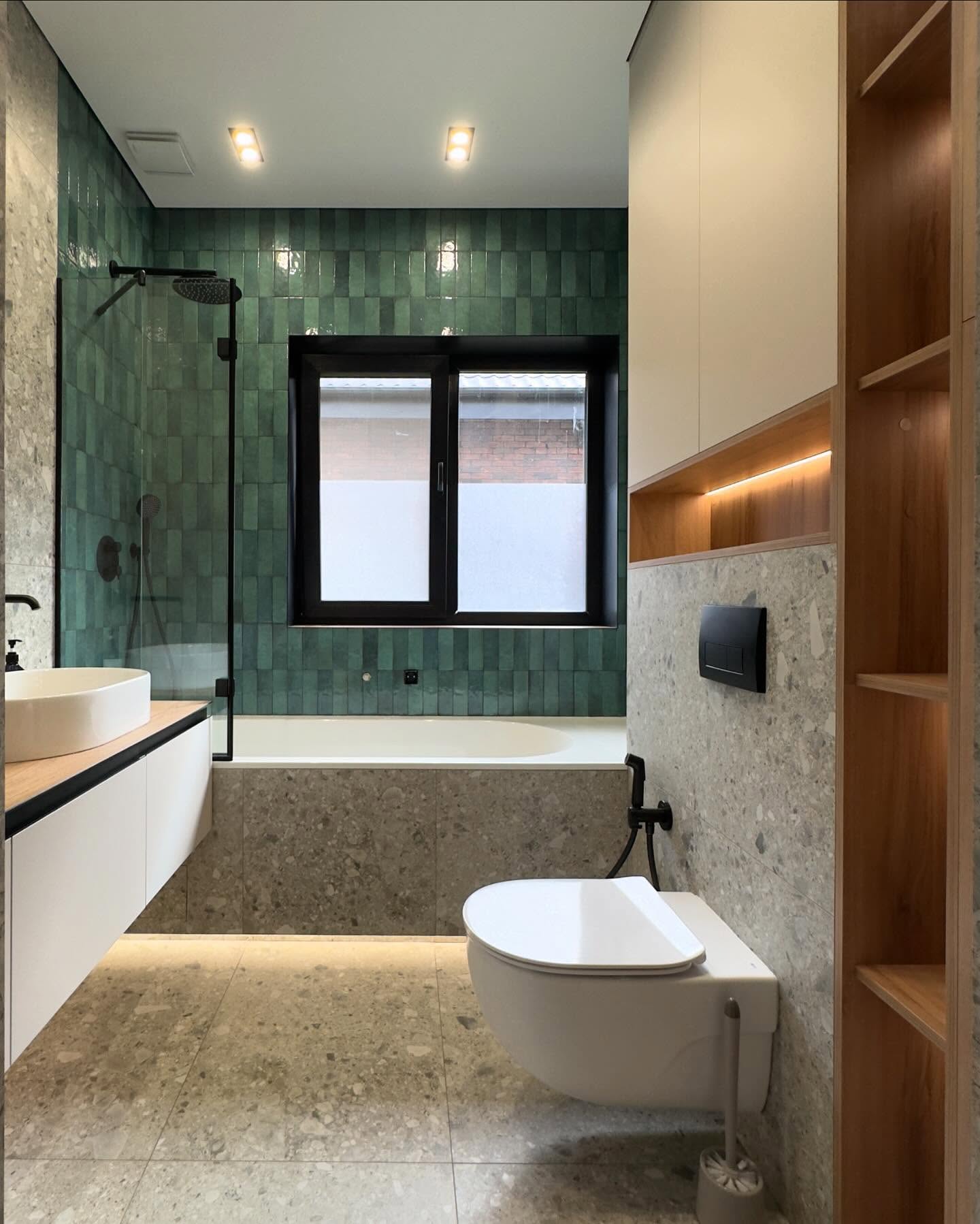 Інтер'єр ванної кімнати у повний ріст: підвісний унітаз, біла підвісна тумба з раковиною, вбудована ванна з зеленою плиткою та вікном, високі дерев'яні стелажі з підсвічуванням.